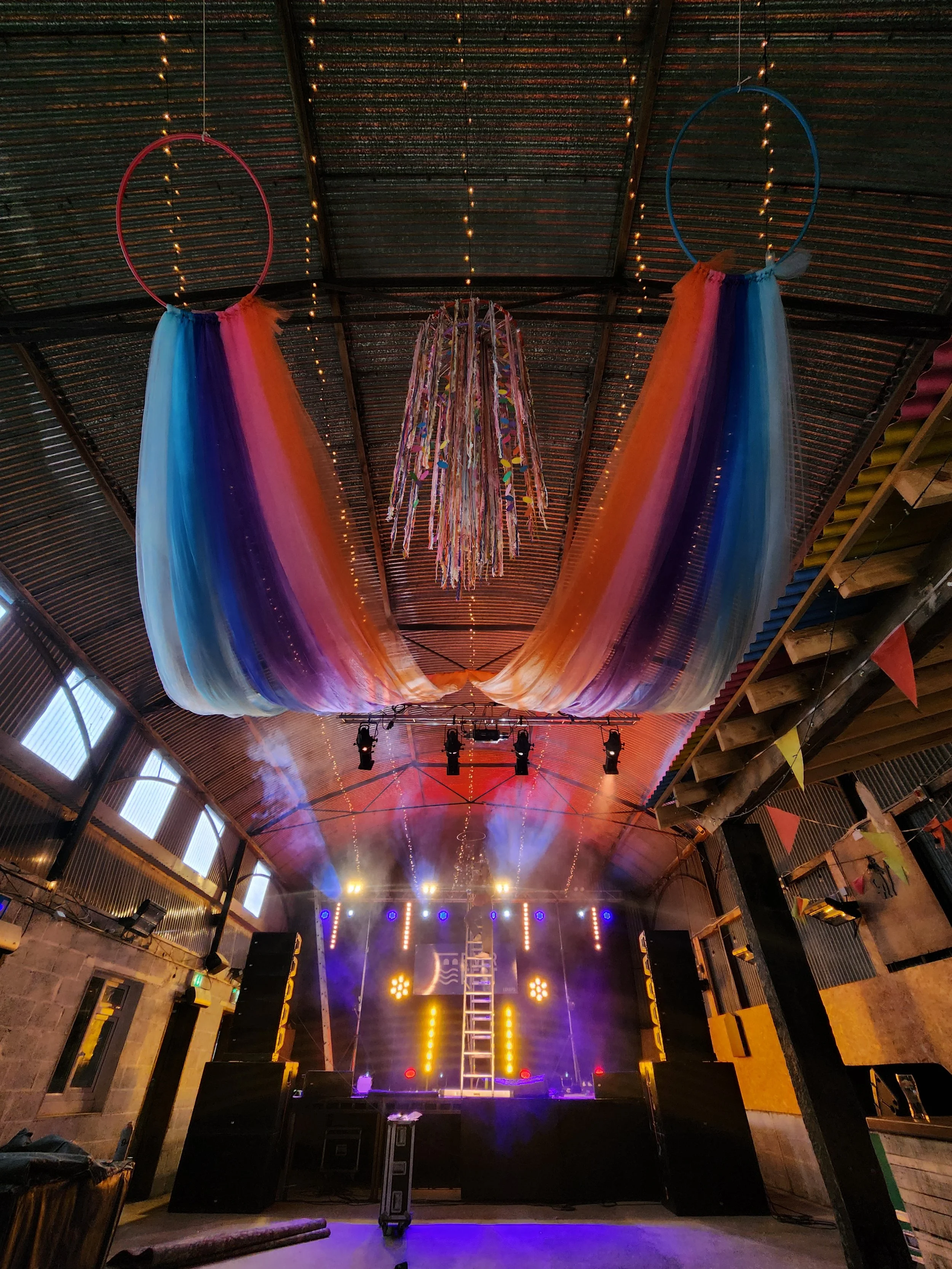 Decorative fabric curtains in rainbow colors hanging from the ceiling over a stage with lighting and smoke effects, set up for an event or concert.