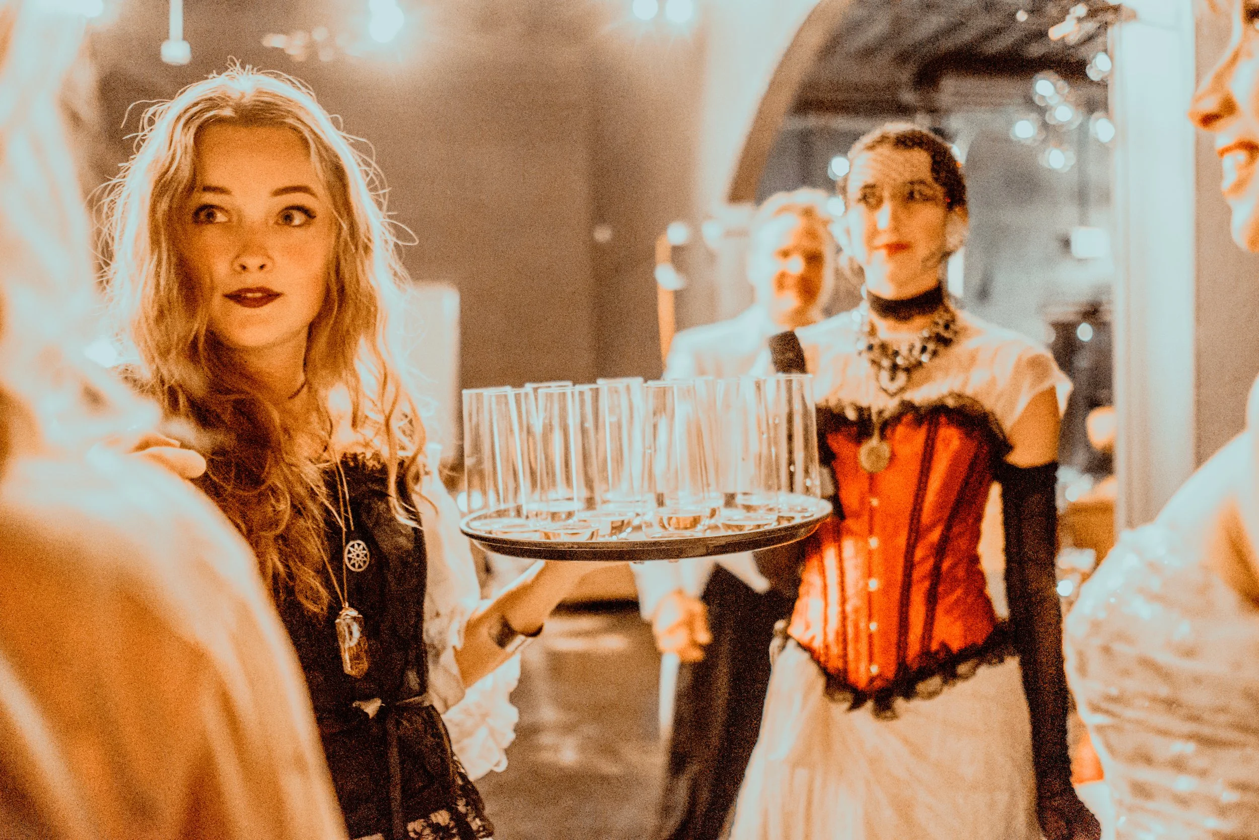 A woman dressed as a goth waitress holding a tray with empty shot glasses in a dimly lit, vintage-style room with others in period or costumes.