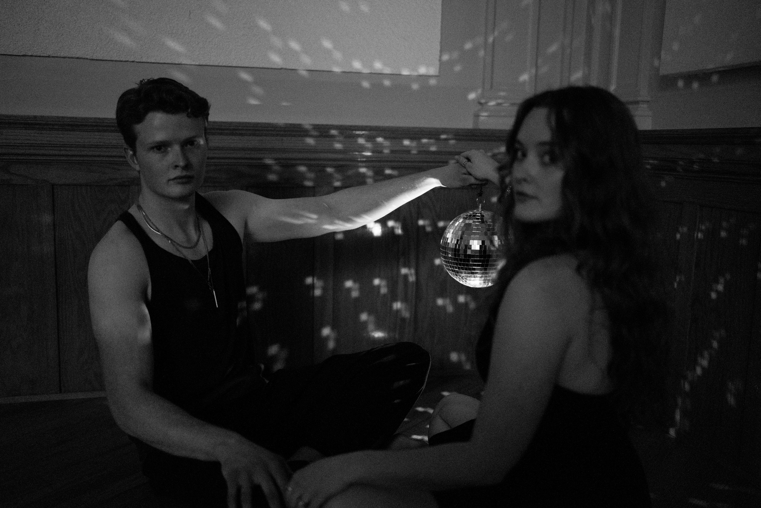A black and white photo of a young man and woman sitting on a floor in a dimly lit room with a disco ball hanging from the ceiling. The young man wears a sleeveless top and has short hair, while the woman has long wavy hair and is dressed in a black 