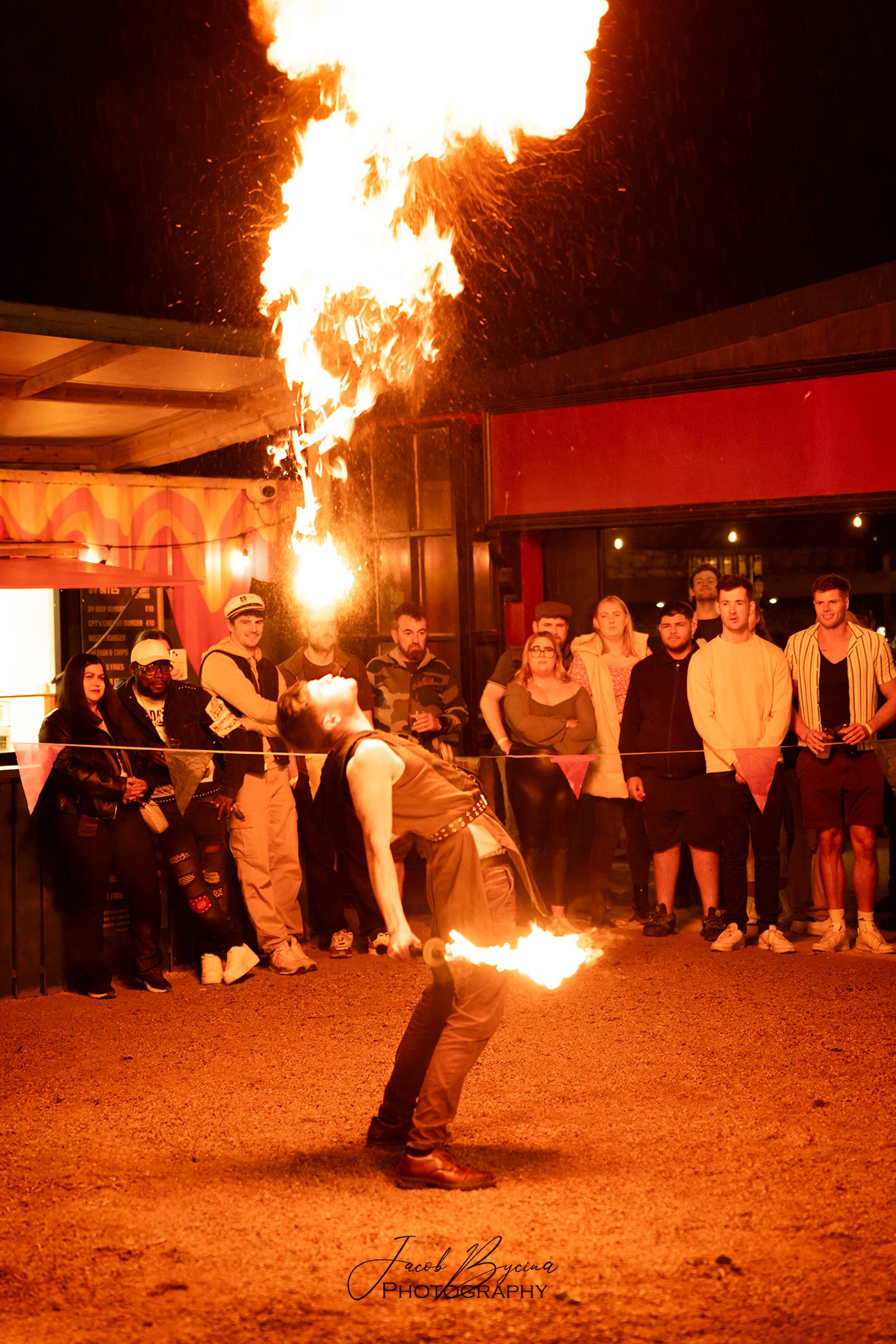 A performer in a sleeveless shirt and dark pants is doing fire breathing or fire dancing outdoors at night, with flames shooting into the air and an audience watching behind a barrier.