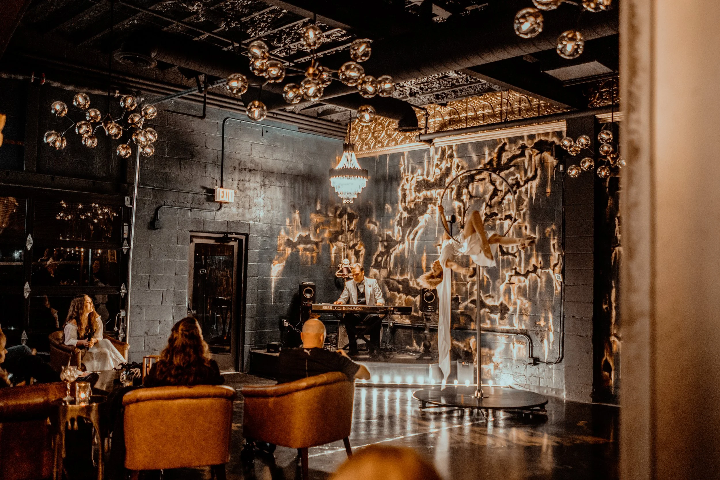Interior of a dimly lit live music venue with a small stage. A musician in a suit is playing a keyboard, and a woman is performing aerial silks on a hoop. Audience members are seated on leather chairs, watching the performance. There are modern light