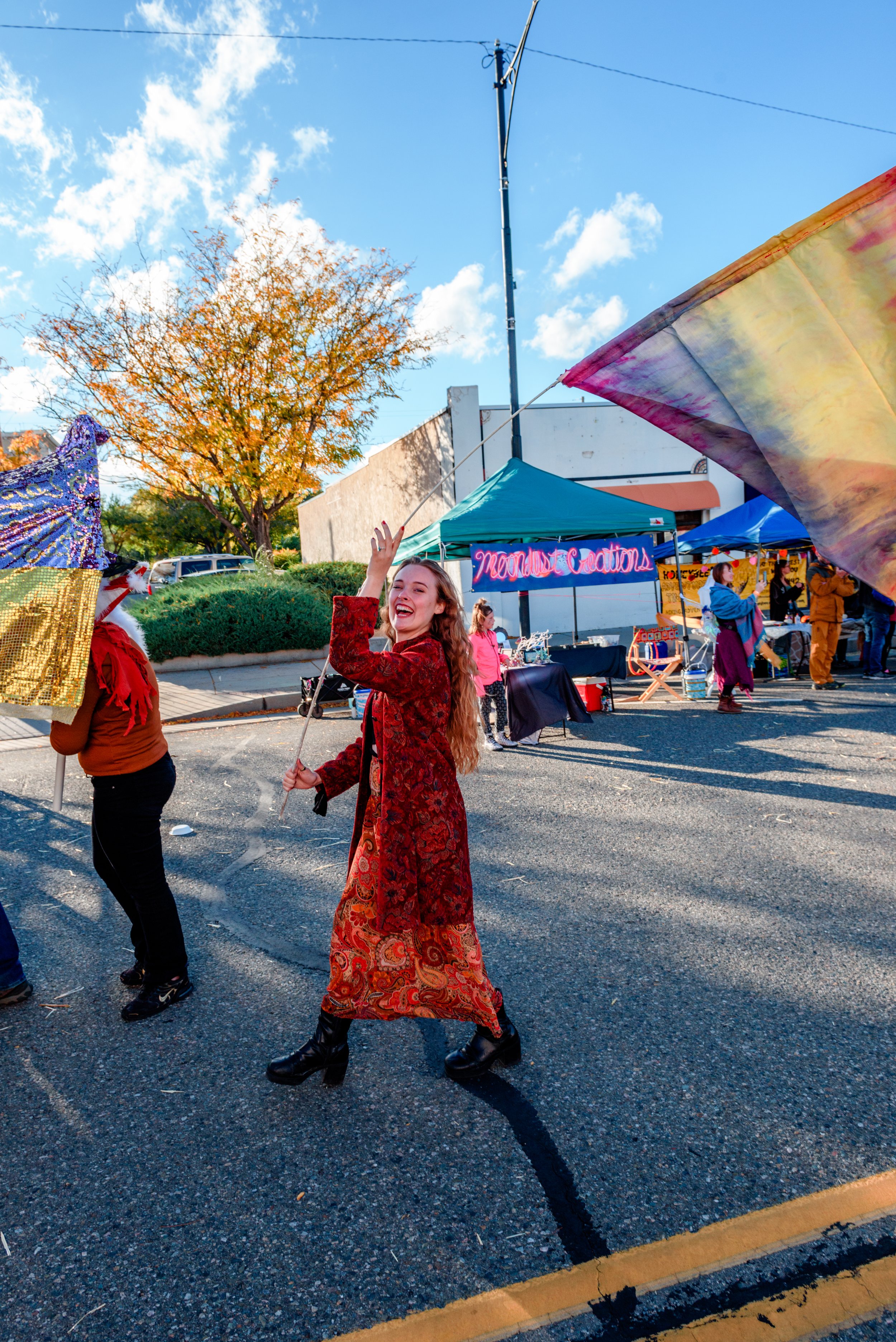 A woman in a long red patterned dress and black boots is smiling and waving a rainbow tie-dye flag at an outdoor festival on a sunny day. There are tents and booths in the background, with a neon sign that reads 'Meow Creations' and trees with fall f