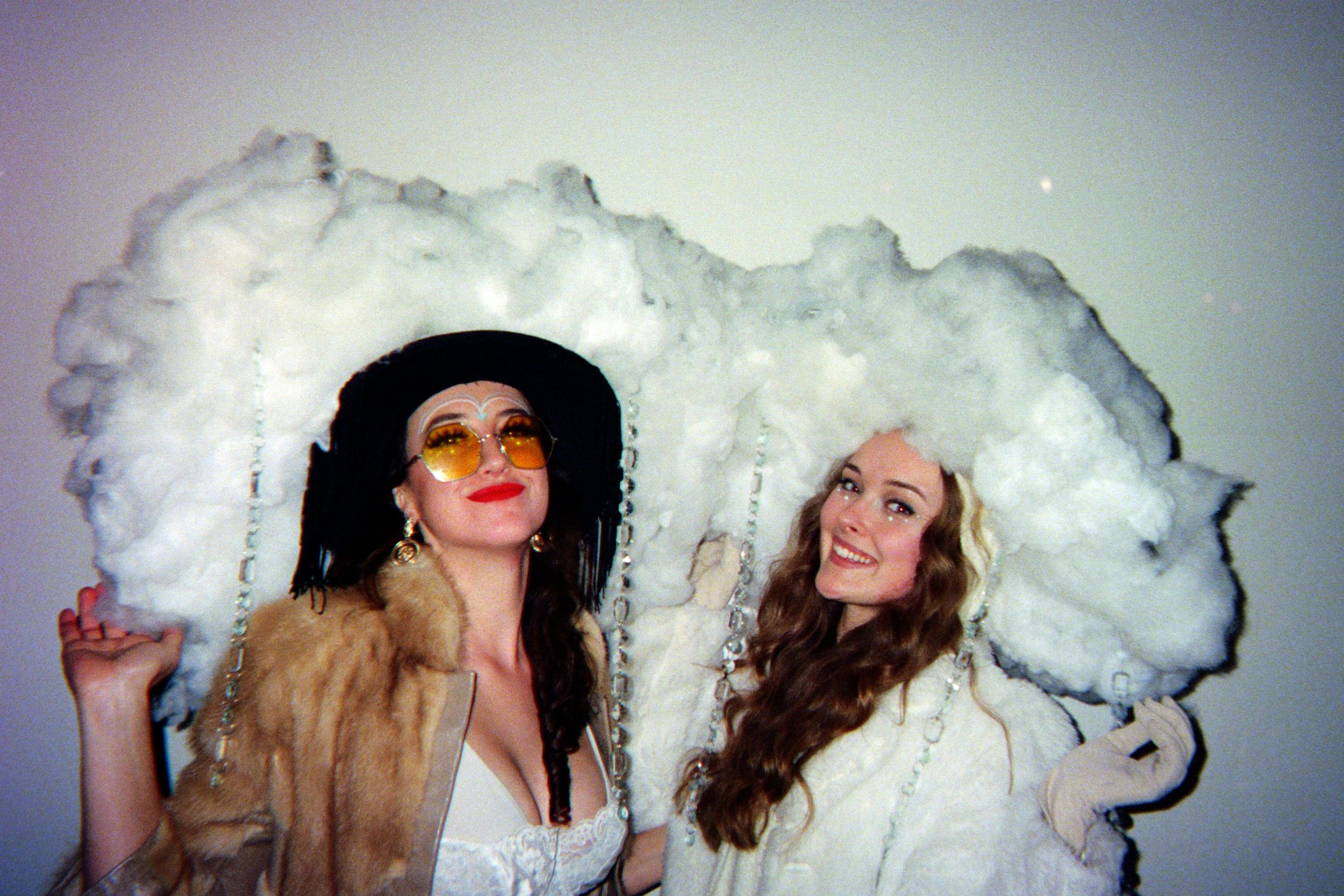 Two women in elaborate costumes. One wears a large black hat, yellow sunglasses, and a fur coat. The other has long curly hair and a white outfit, standing under a cloud-like prop.