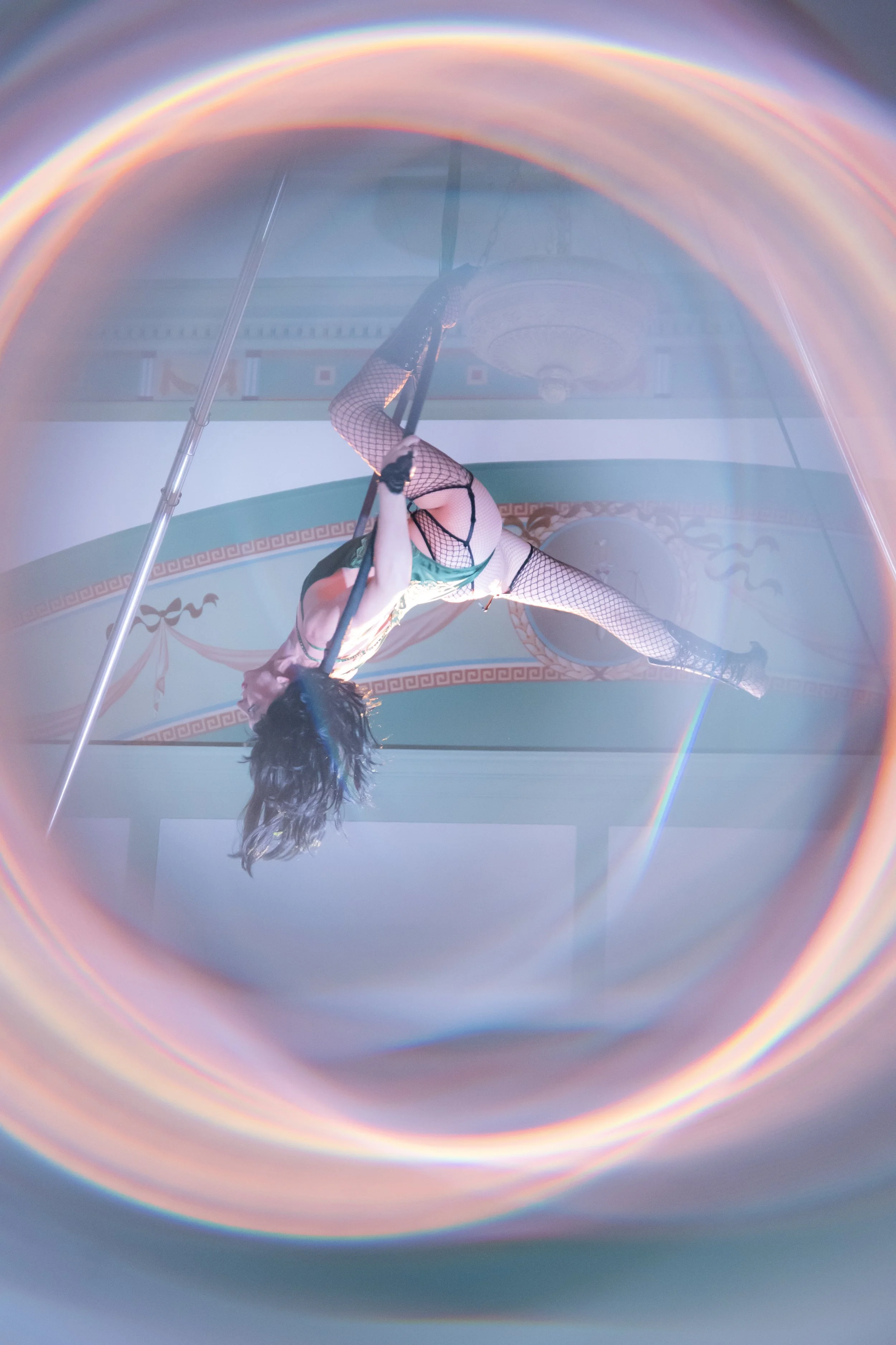 A performer in fishnet stockings and a costume performing an aerial dance on a hoop, viewed through a circular object with a rainbow-colored edge, in a decorated room.