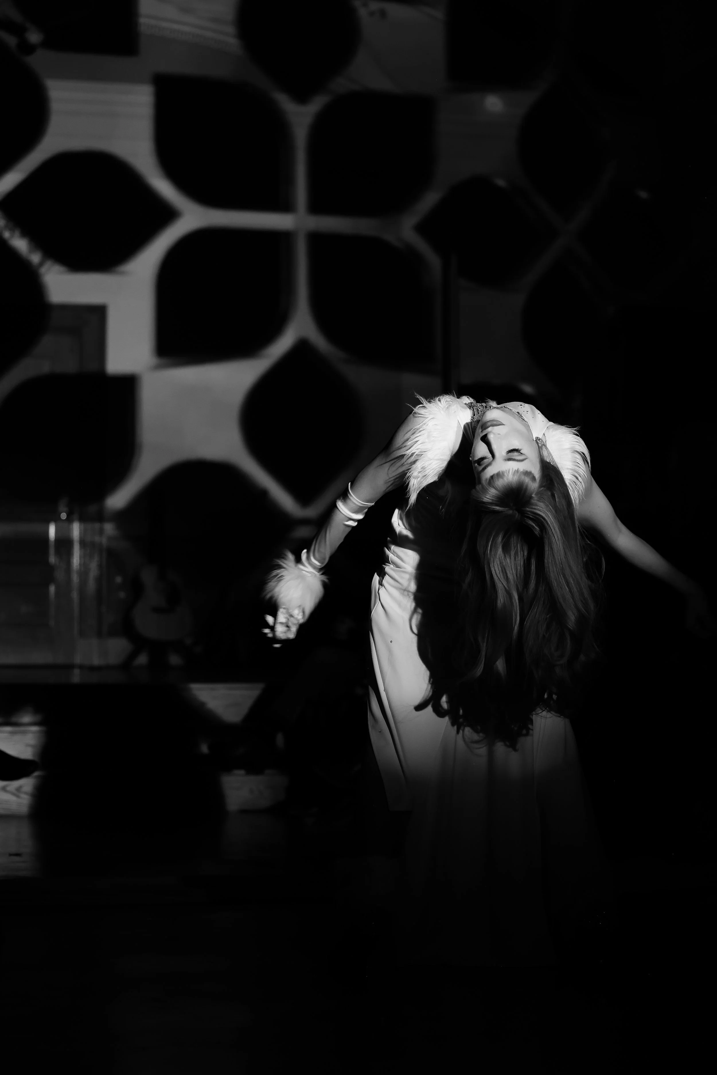 A woman dancing on stage, bending backward with her long hair hanging down, illuminated by spotlight in a dark room alongside an abstract background.