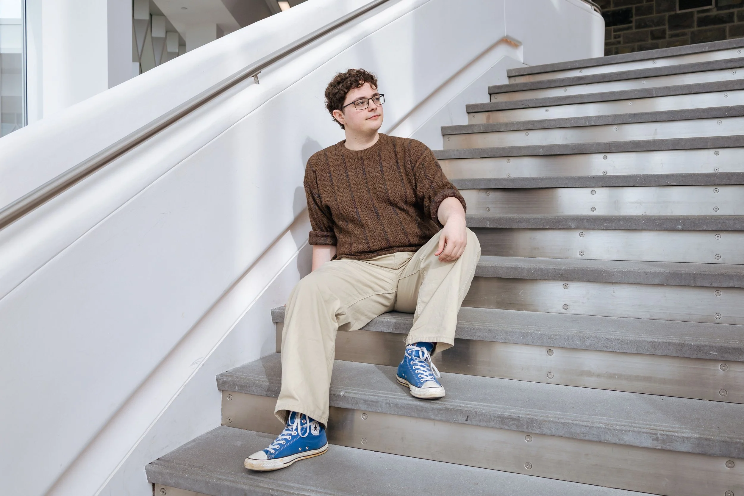 A young person (Brian Bush) with curly hair, glasses, wearing a brown sweater, beige pants, and blue sneakers, sitting on indoor stairs with a white railing.