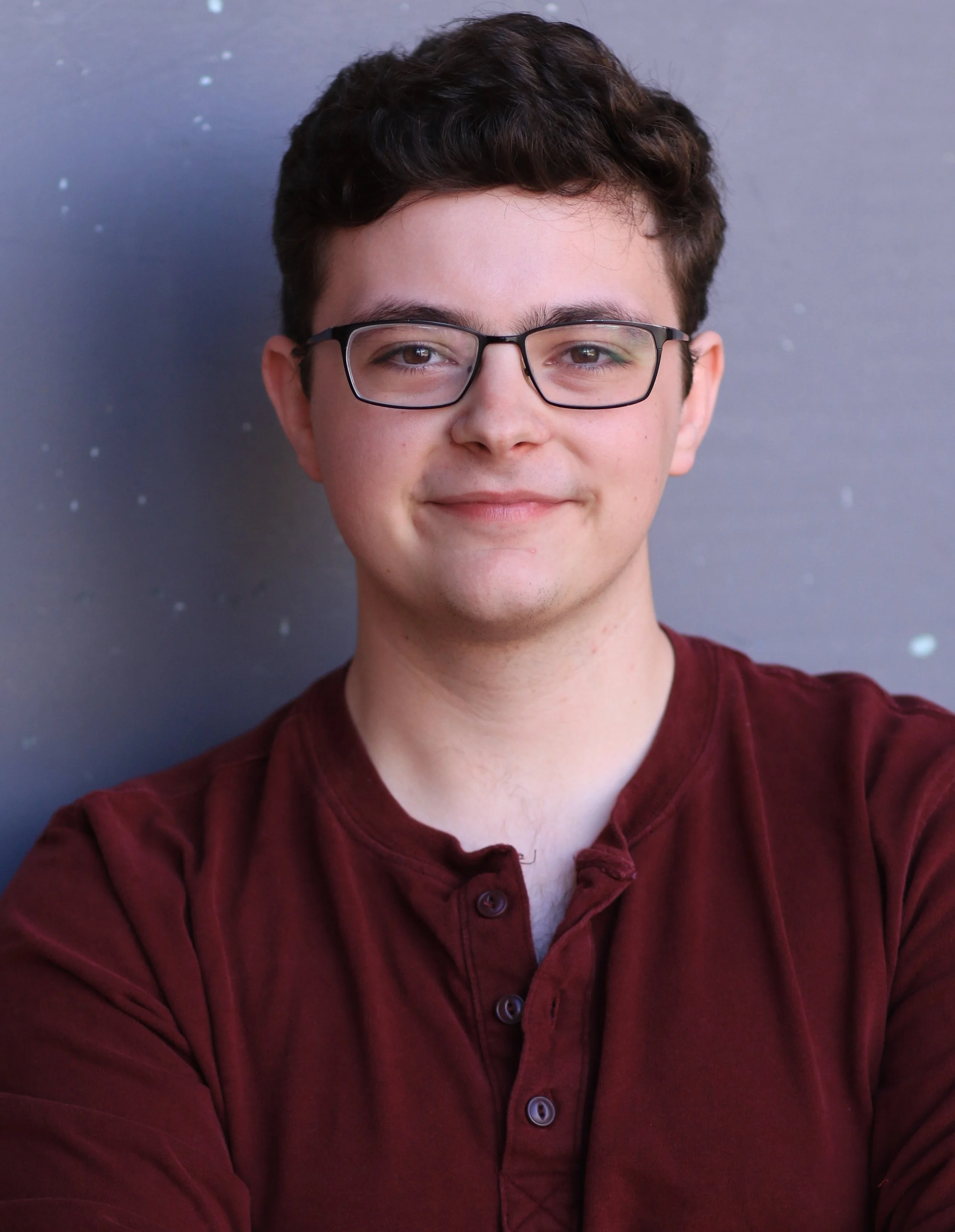A young man (Brian Bush) with brown hair, glasses, and a slight smile, wearing a maroon shirt, standing against a gray wall.