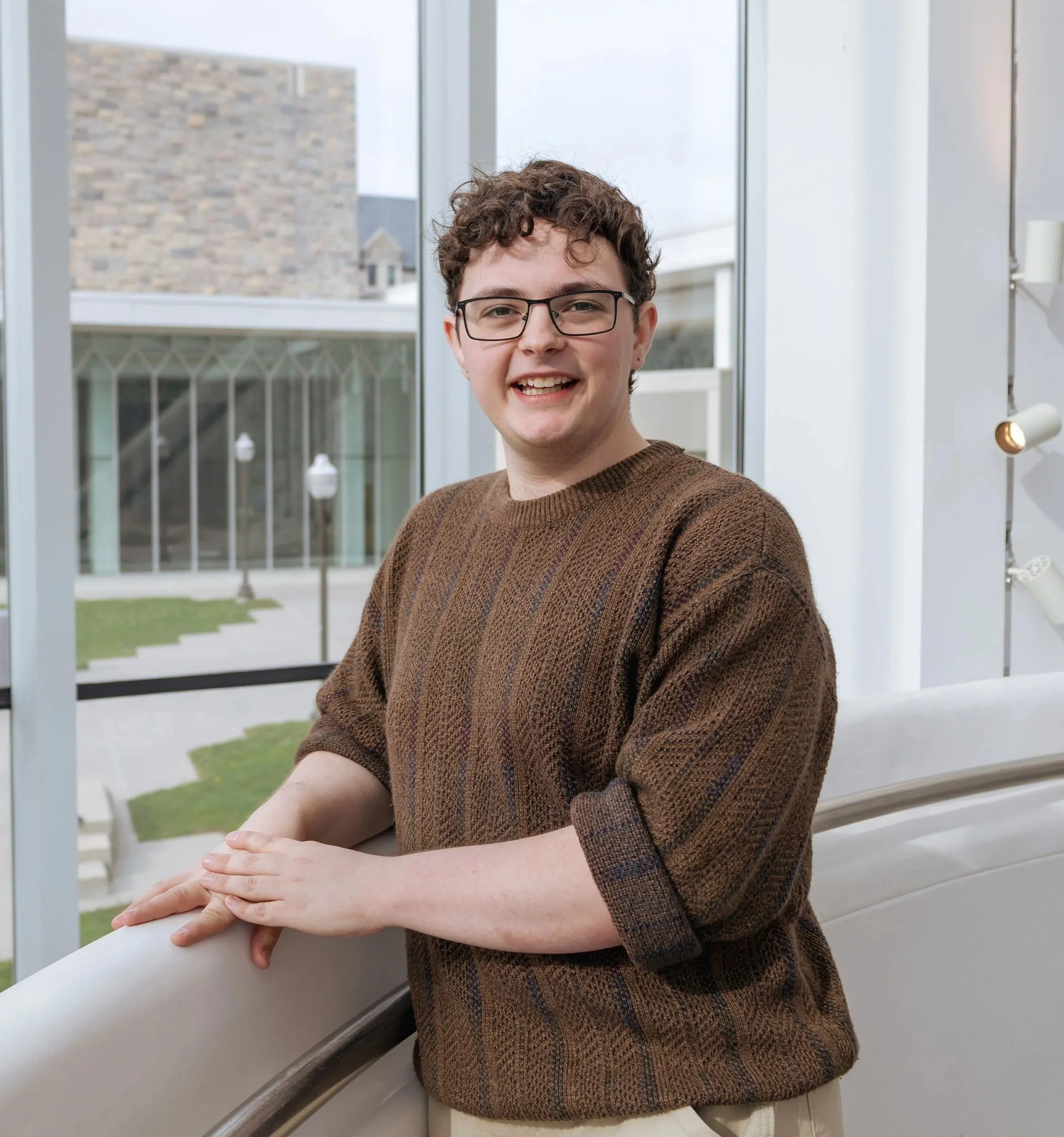 Young man (Brian Bush) with curly hair and glasses smiling, standing indoors near a railing with large windows behind him showing a building and outdoor walkway.