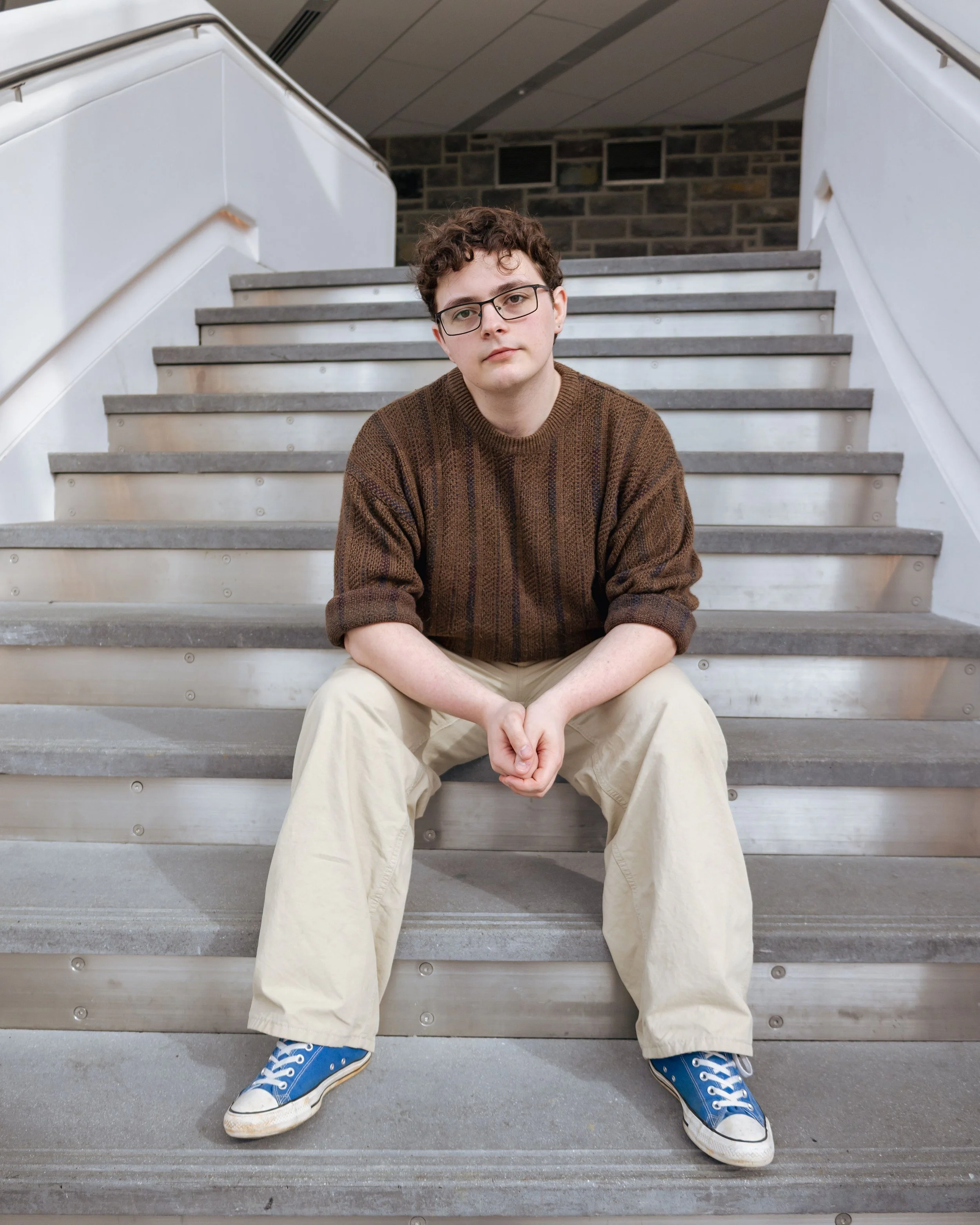 A young man (Brian Bush) with curly hair and glasses sitting on a staircase, wearing a brown sweater, beige pants, and blue sneakers, looking at the camera.
