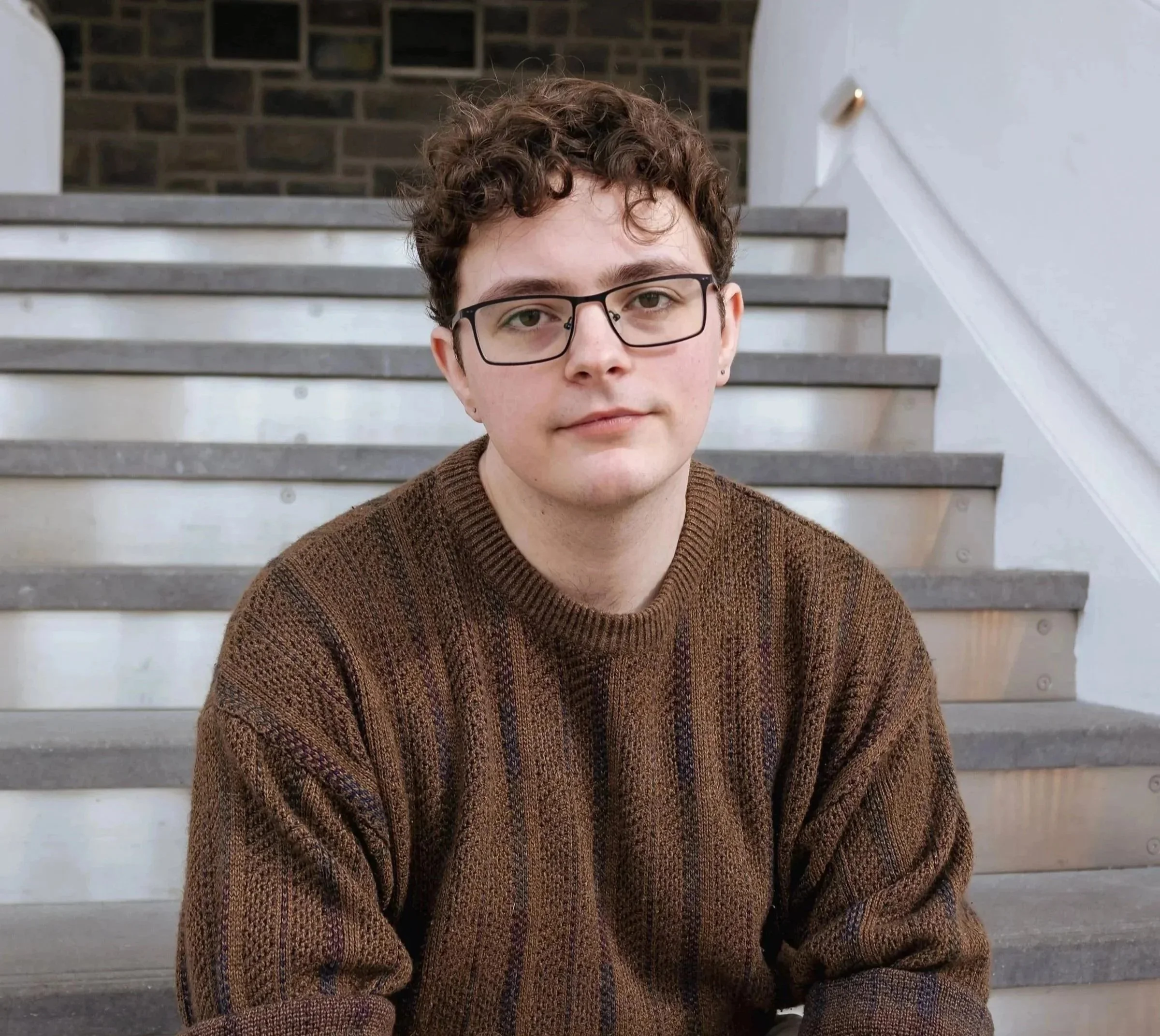 Young man (Brian Bush) with curly hair and glasses sitting on stairs, wearing a brown sweater.