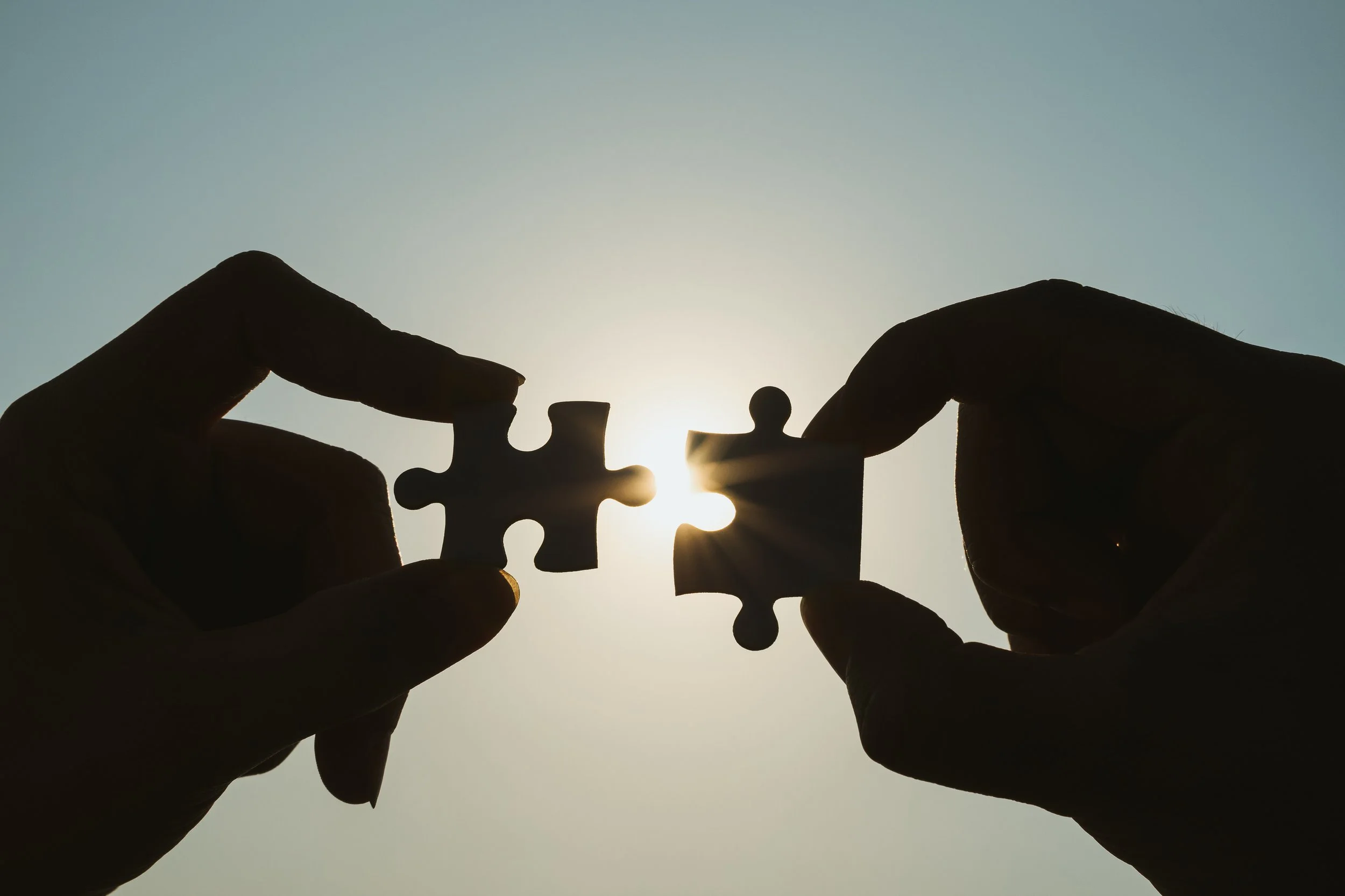 Two hands each holding a puzzle piece, fitting them together in front of a bright sky with the sun shining through, symbolizing problem-solving or connection.