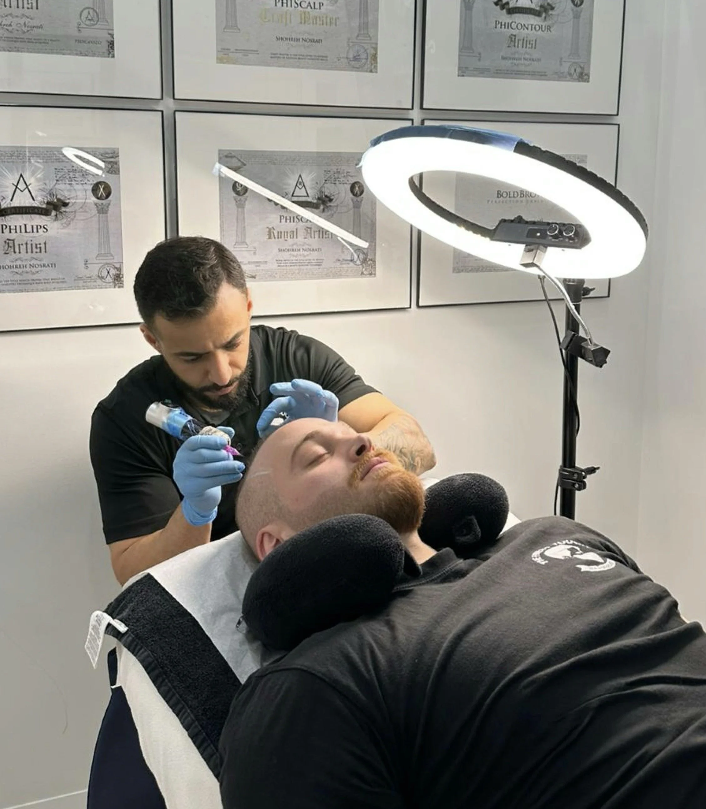 Tattoo artist performing a tattoo on a man's arm in a tattoo studio. The studio has framed certificates and diploma posters on the wall and a large ring light on a stand illuminating the work area.