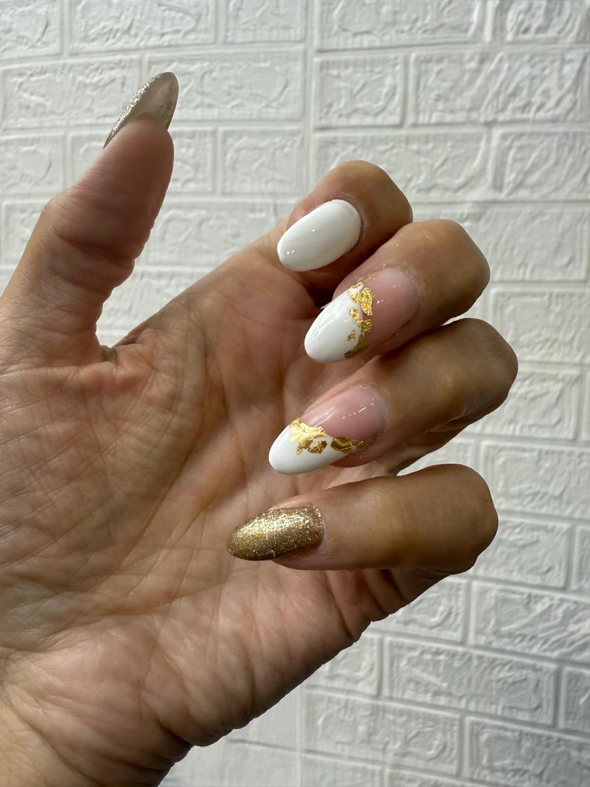 Hand with decorated nails, featuring white, clear with gold accents, and gold glitter designs, held against a textured white brick wall background.