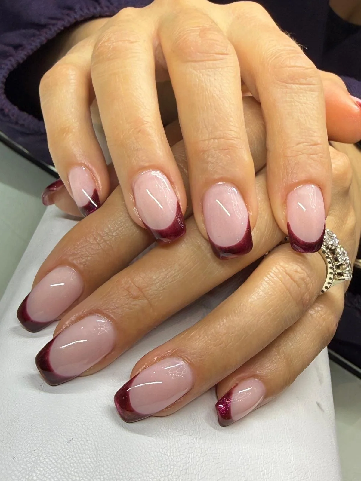 Close-up of manicured hands with French tips and red-edged nail polish, rings on fingers, on a white surface.