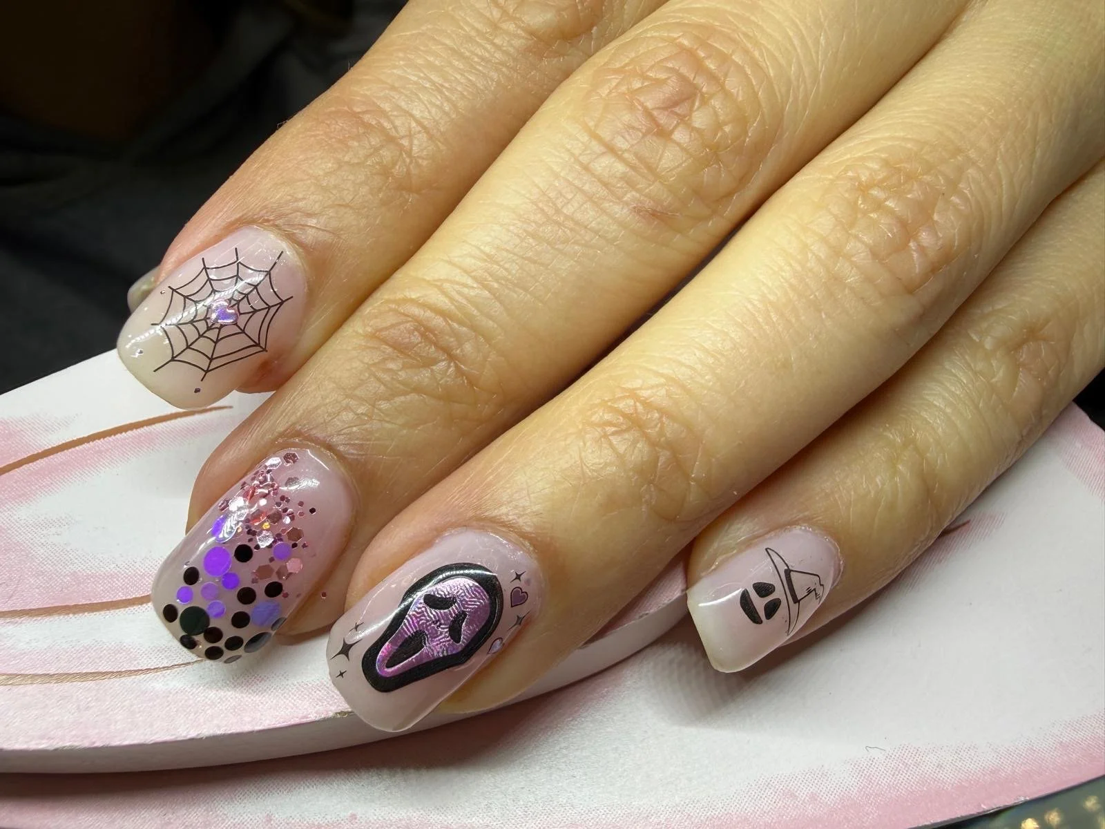 Close-up of a hand with decorated fingernails featuring Halloween-themed designs like a spider web, a witch's hat, and glitter.
