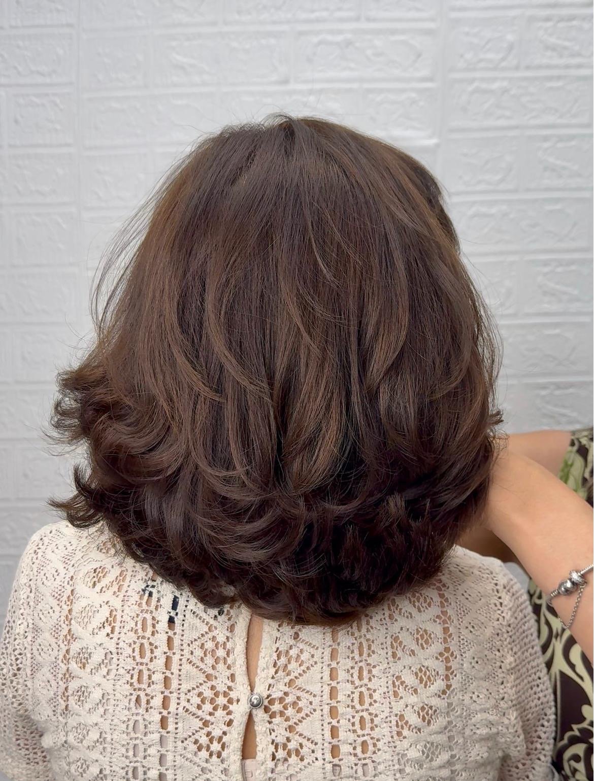 A woman with shoulder-length, layered, wavy brown hair, wearing a white lace top with a keyhole button detail, sitting against a white brick wall.