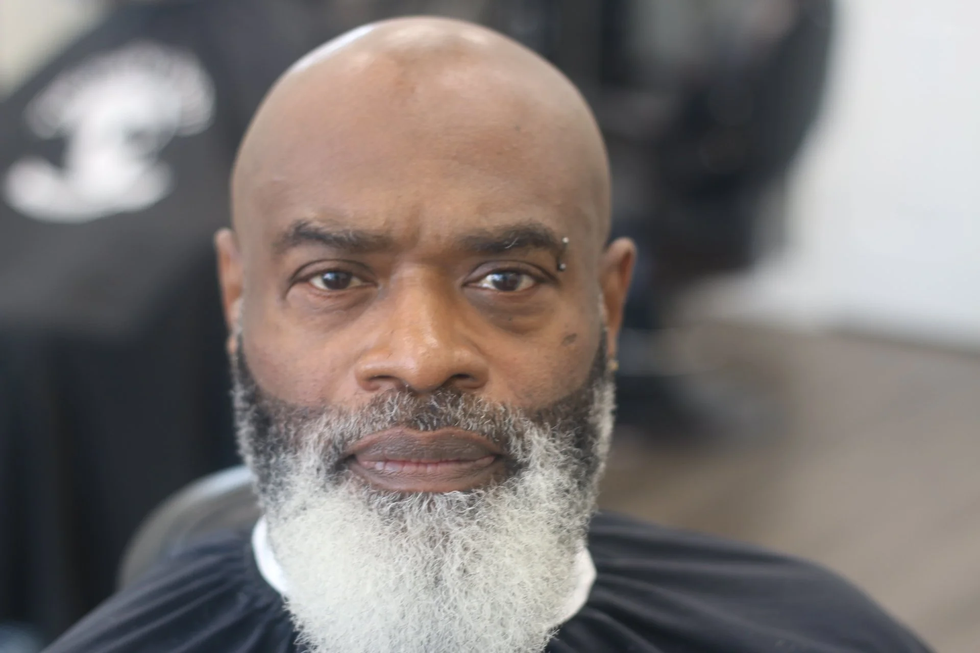 Close-up of a calm African American man with a bald head, a bearded face with gray hair, and brown eyes, sitting on a black chair in what appears to be a salon or barbershop.