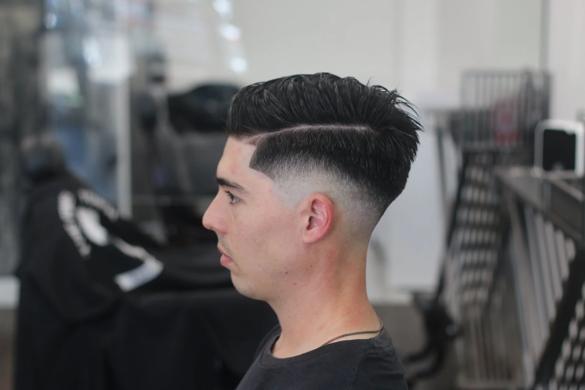 Side profile of a young man with slicked-back hair and a fresh fade haircut, sitting in a barbershop.