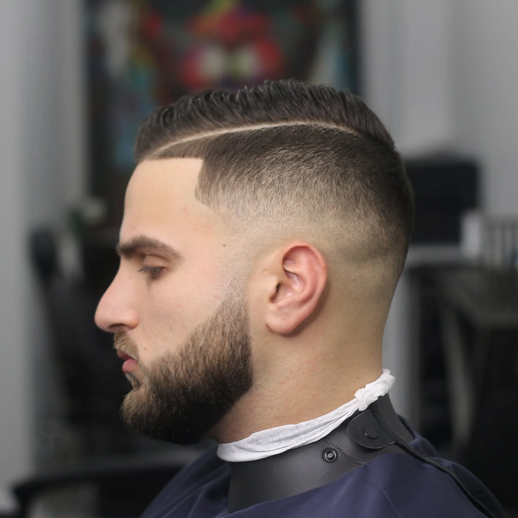 A man with a well-groomed hairstyle in a barbershop, with a high fade haircut and a side part, wearing a barber cape and a white neck strip.