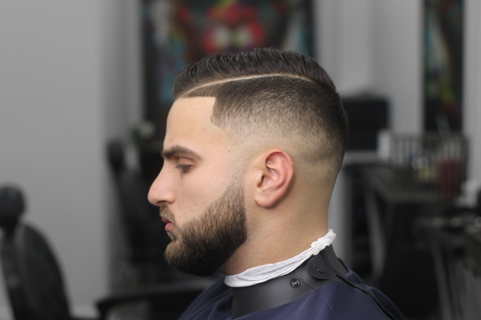 Man with a modern undercut hairstyle, beard, side profile, sitting in a barbershop chair.