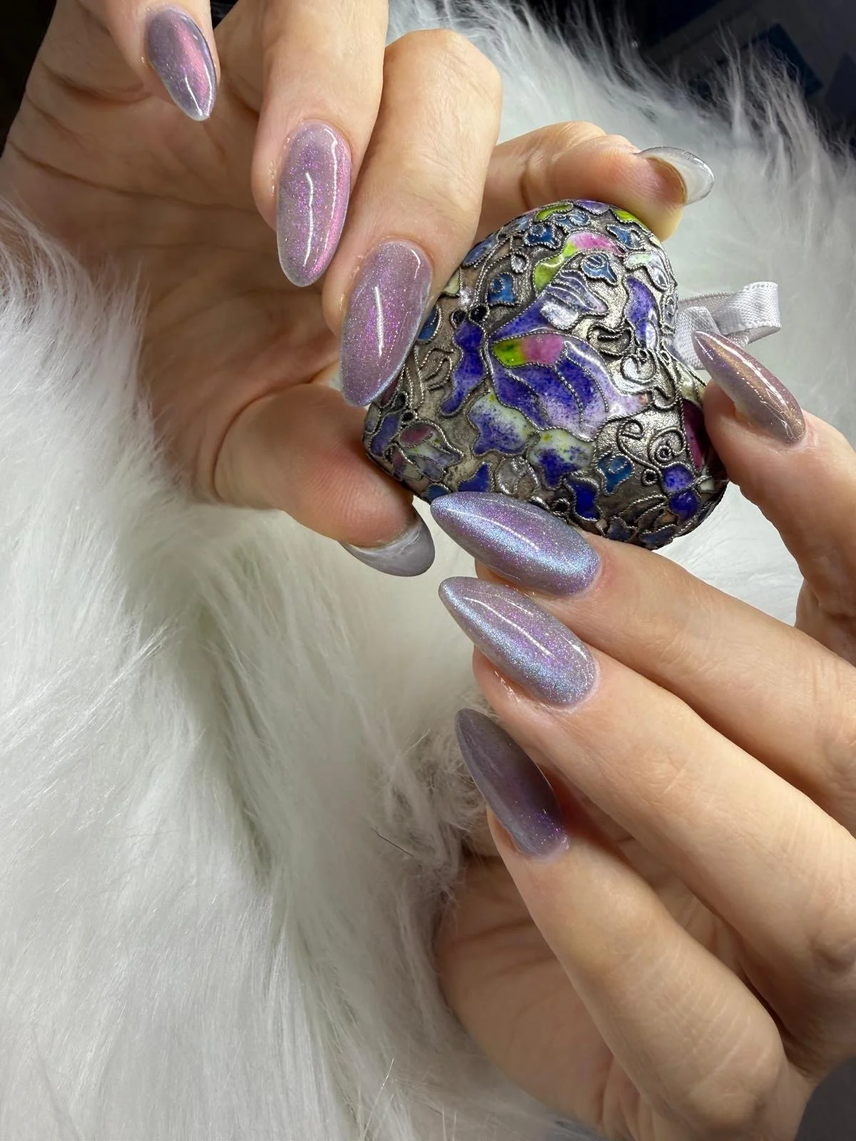 Close-up of hands with holographic, glittery nail polish holding a decorative heart-shaped object with colorful floral motifs, against a white fur background.