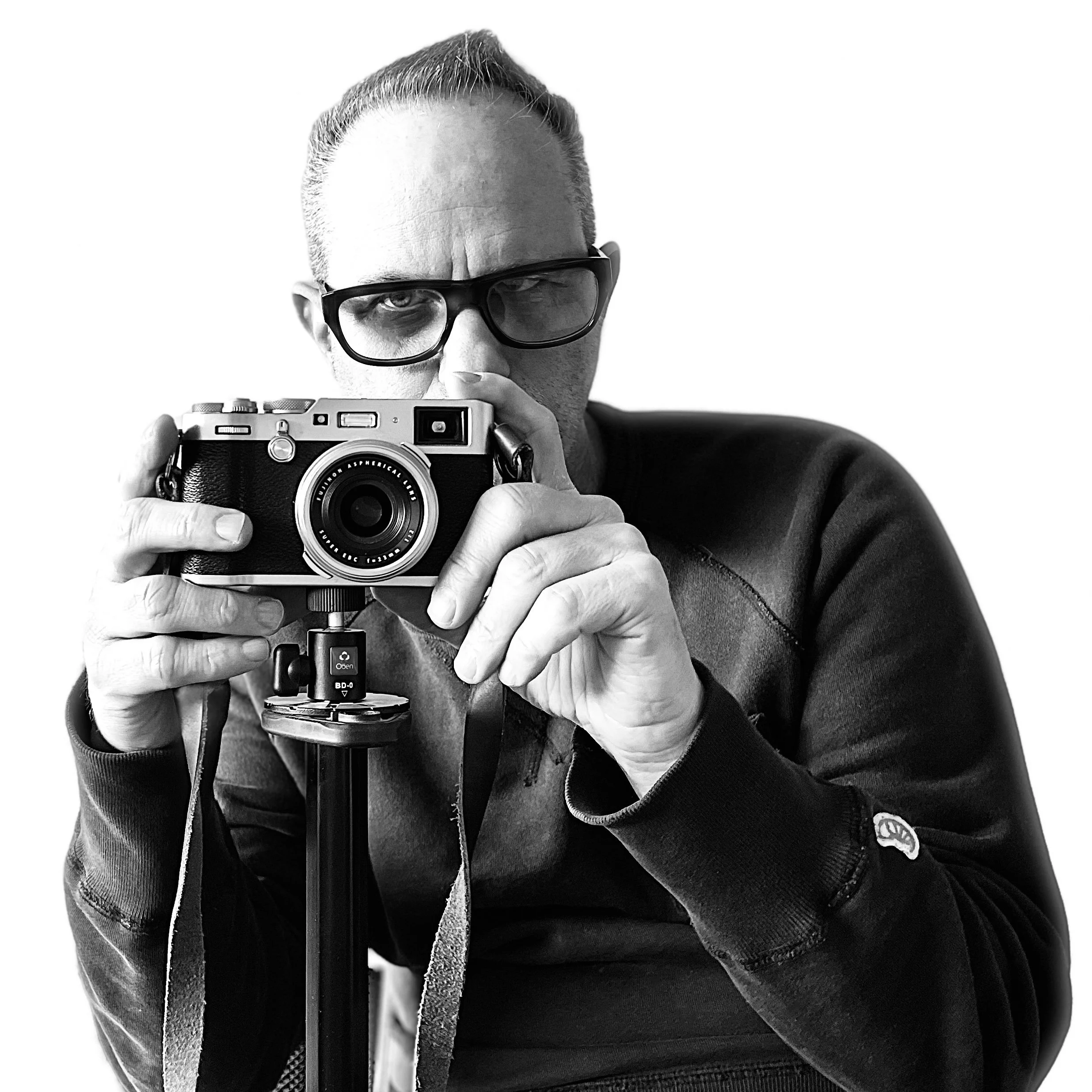 Black and white photo of a man with glasses holding a vintage camera on a tripod, looking directly at the camera.