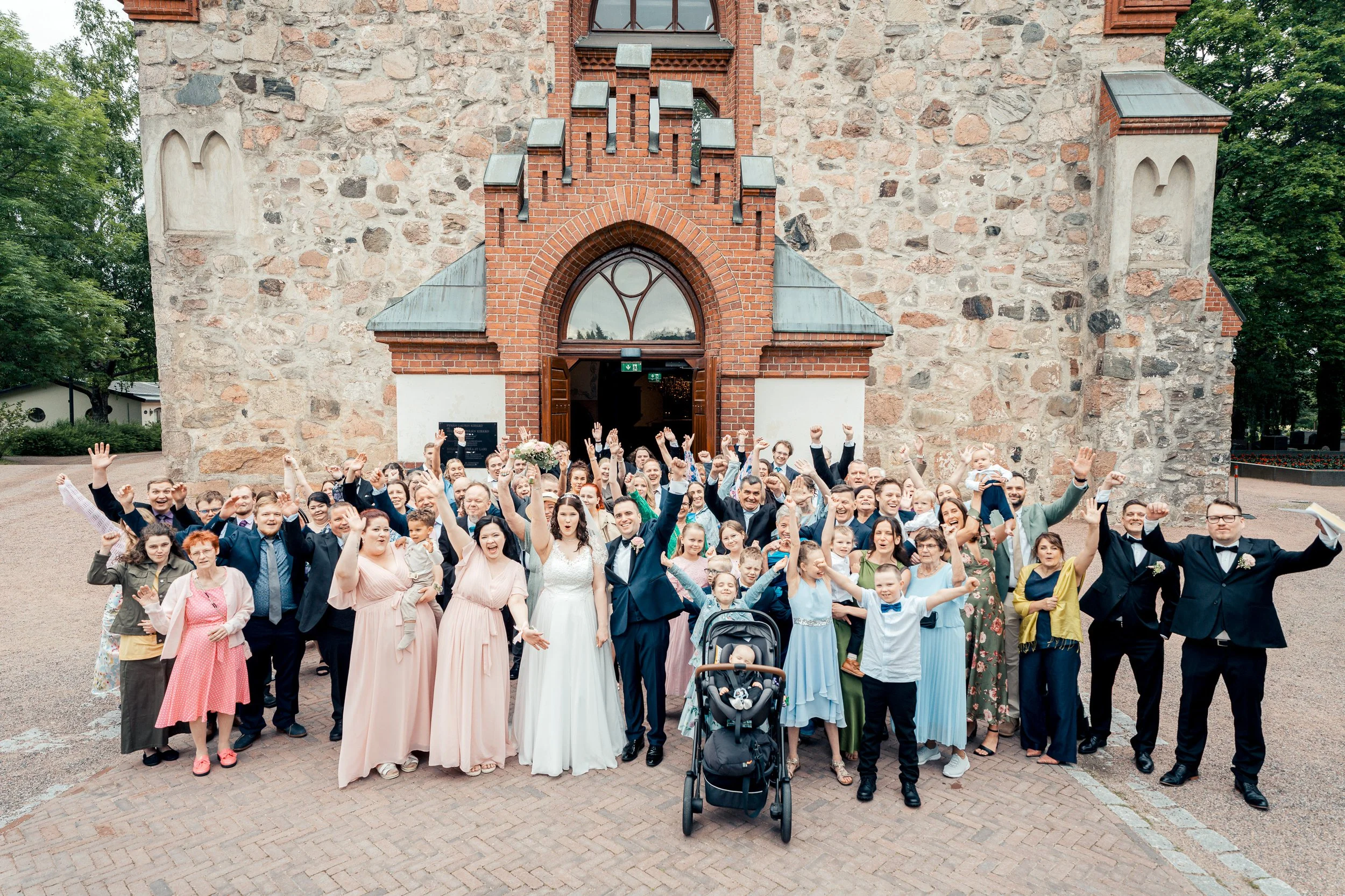 Hääpari ja juhlaväki ryhmäkuvassa Pyhän Laurin kirkon edustalla Vantaalla. Hääkuvaus Vantaa - Hyvän mielen hääkuvaaja Sami Turunen.
