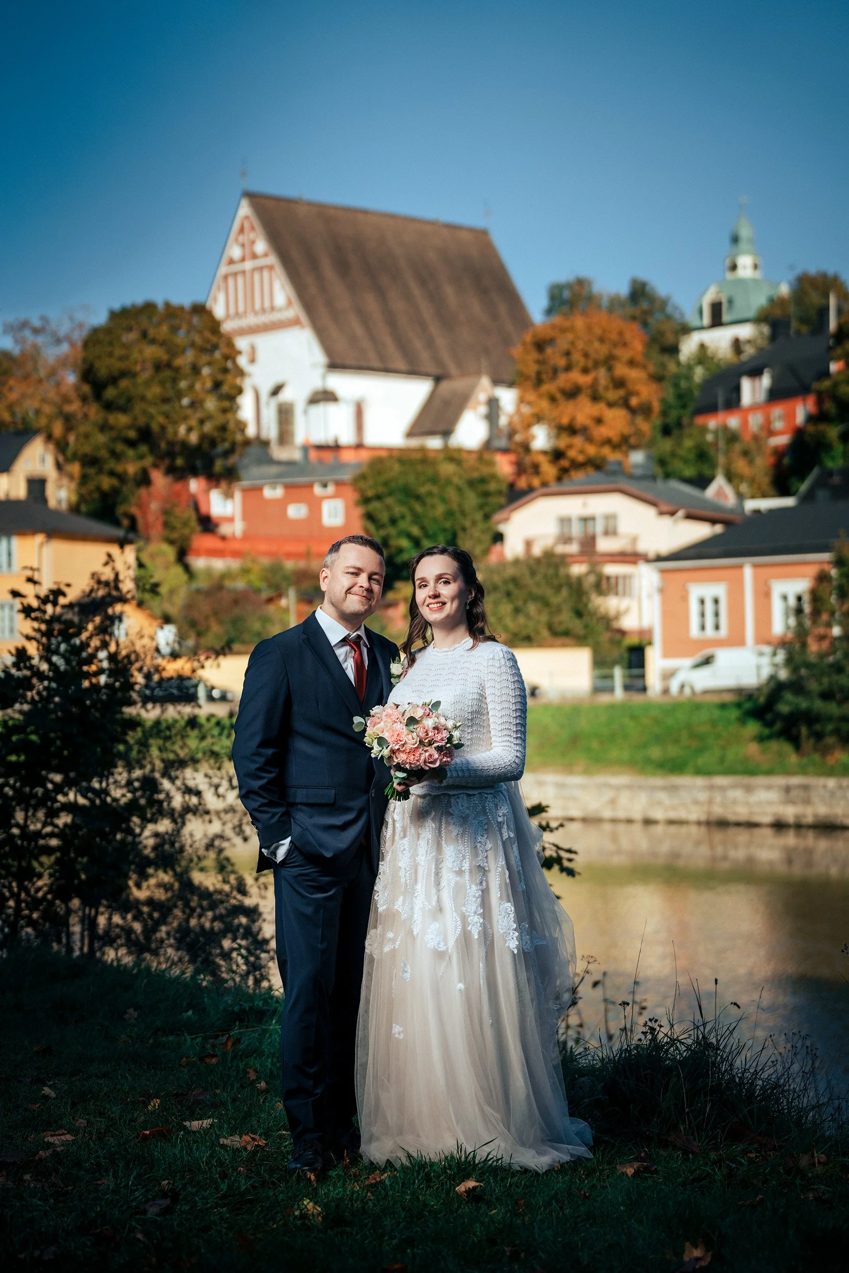 Onnellinen hääpari poseeraa Porvoonjoen rannalla, taustalla näkyy ikoninen Porvoon kirkko. Hääkuvaus Porvoo – hyvän mielen valokuvaaja Sami Turunen.