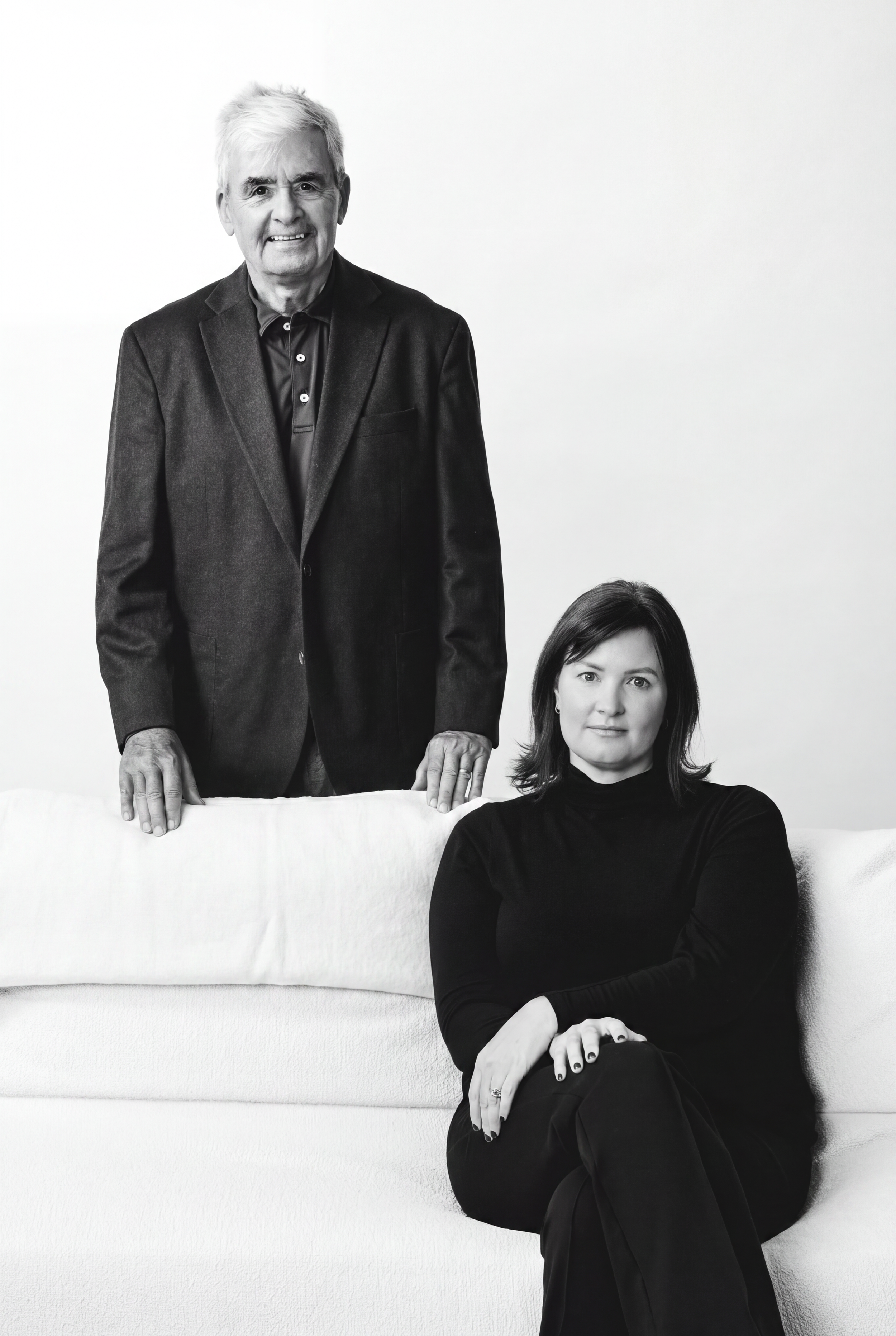Black and white portrait of an elderly man standing behind a seated woman, both dressed in dark clothing, against a plain background.