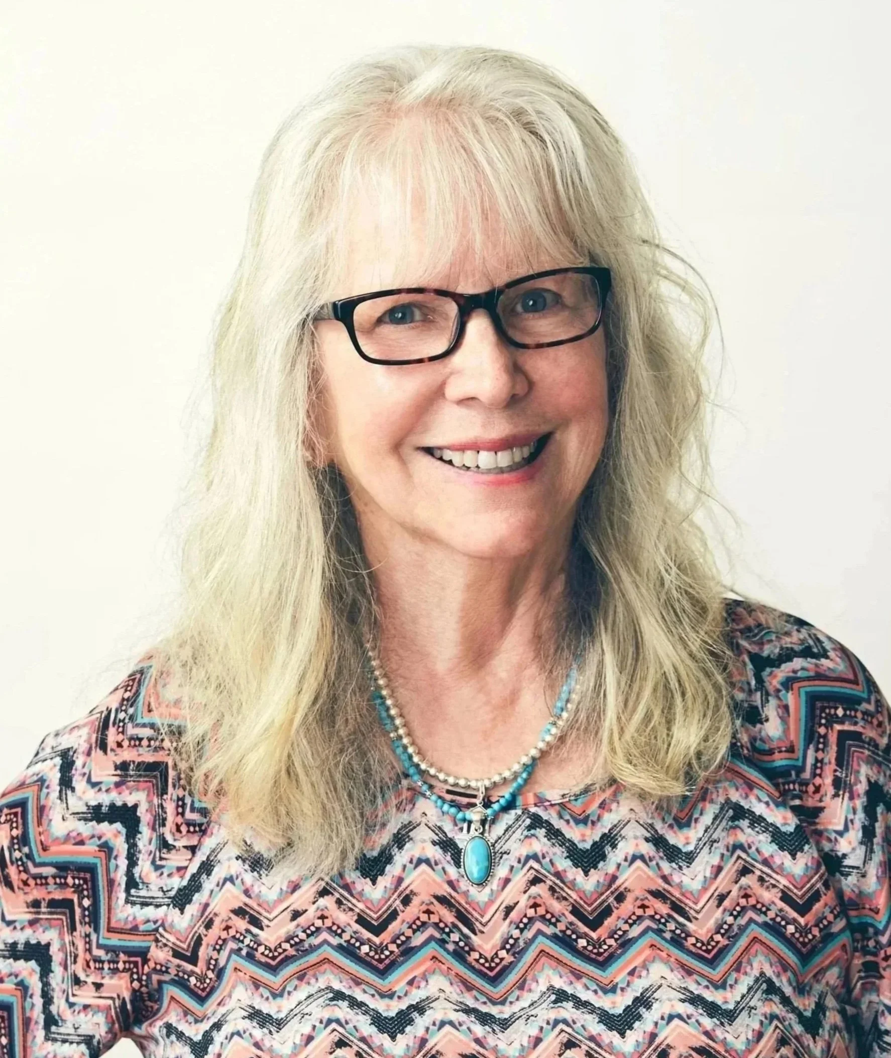 A smiling woman with long gray hair and glasses, wearing a patterned top with pink, turquoise, and black designs, and a turquoise gemstone necklace.