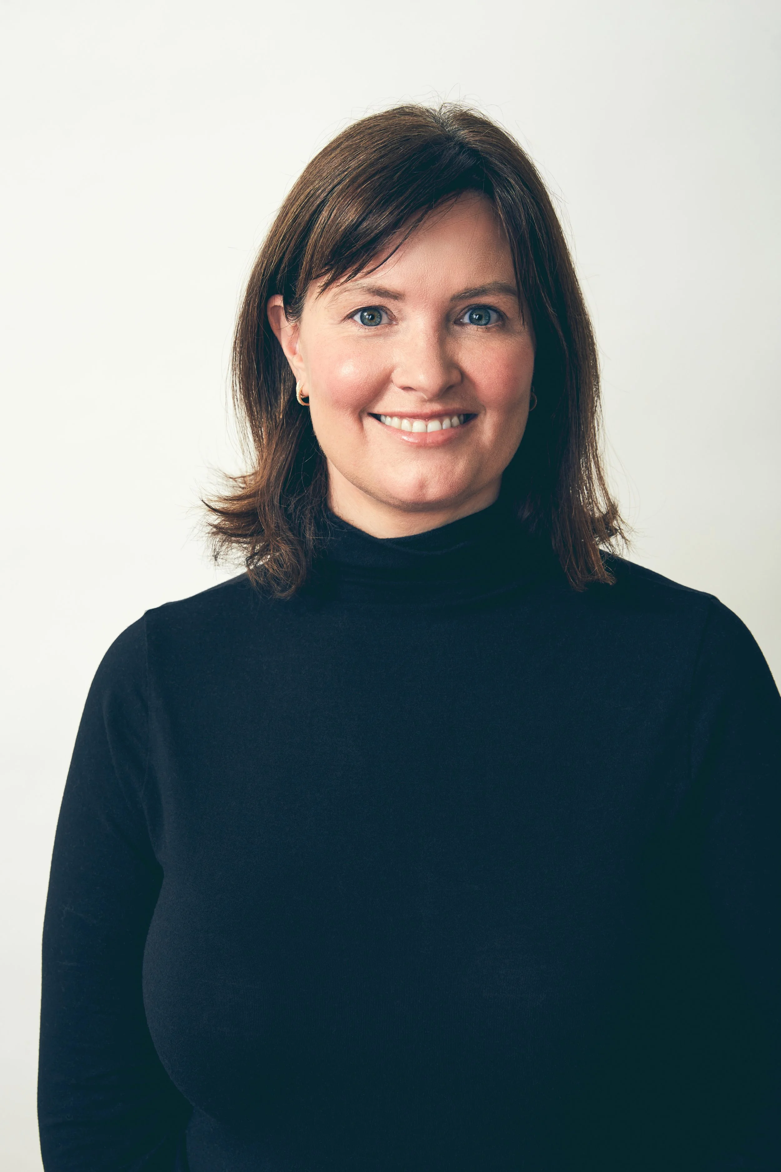 A woman with shoulder-length brown hair, wearing a black turtleneck sweater, smiling and looking directly at the camera.