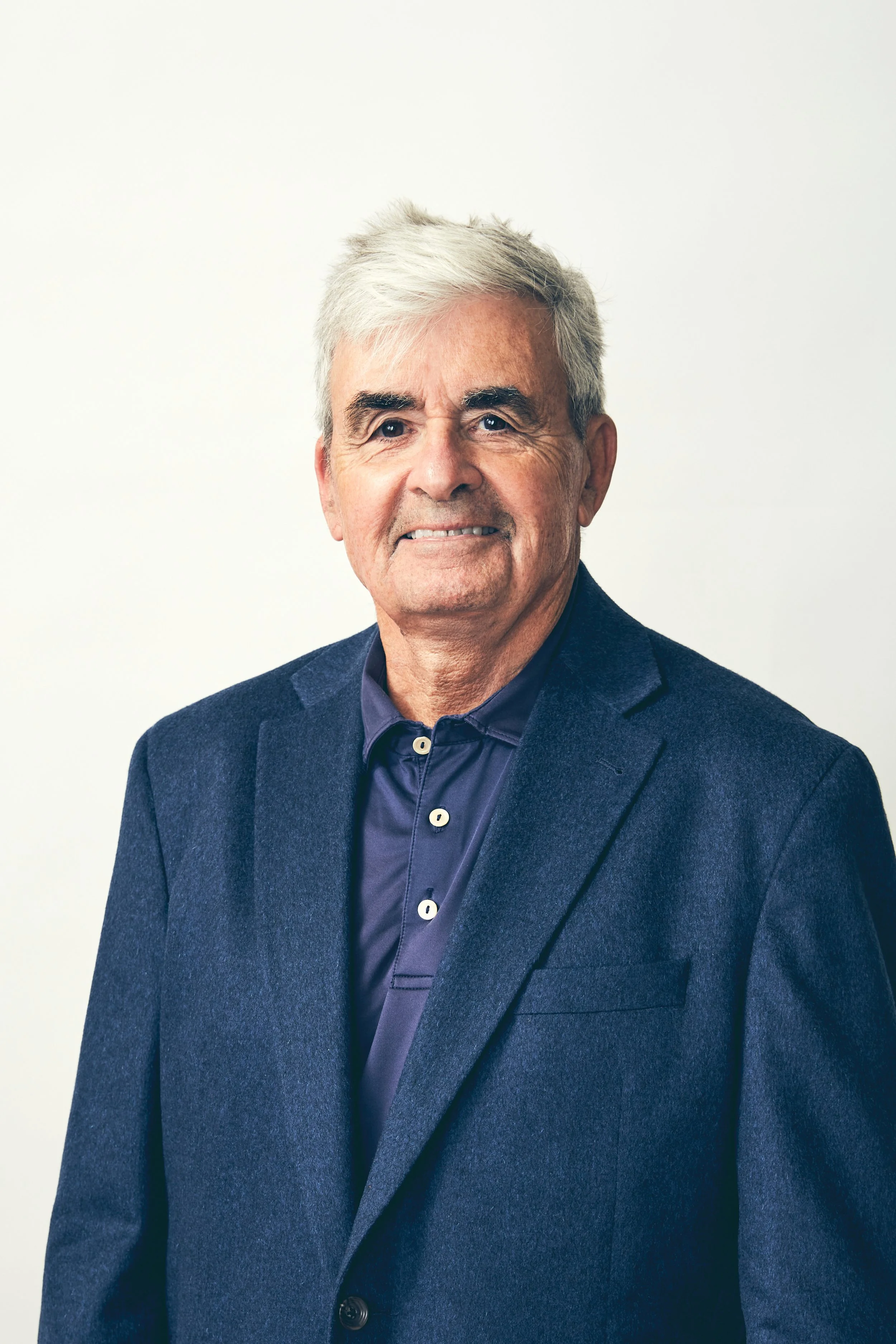 A portrait of an older man with gray hair, wearing a dark blazer over a dark button-up shirt, standing against a light, plain background.