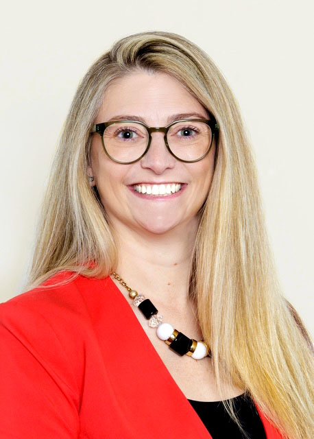 Portrait of a woman with long blonde hair, glasses, and a bright smile, wearing a red blazer and a black top, against a neutral background.