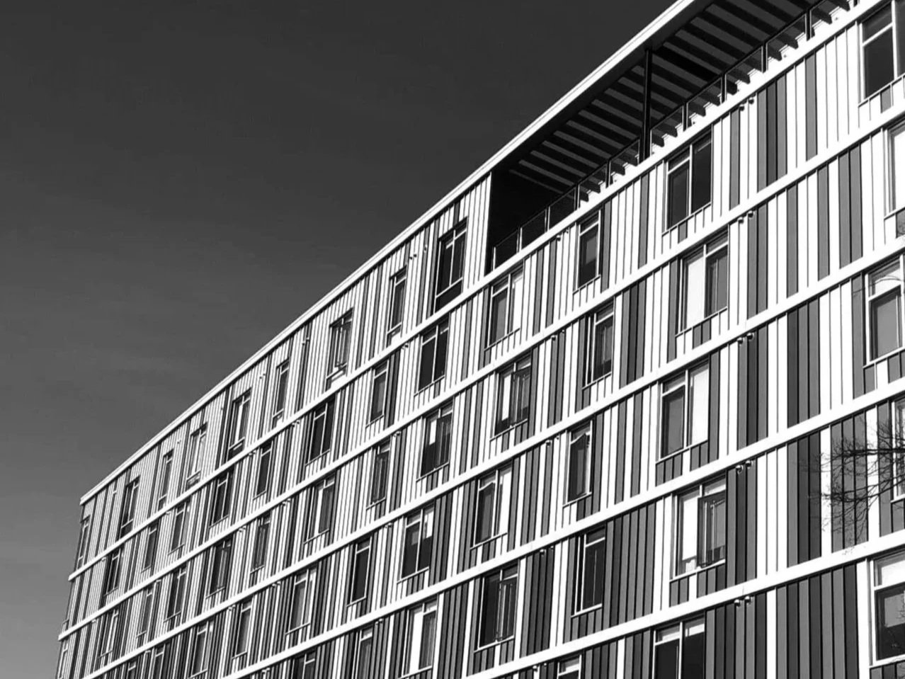 Black and white photo of a modern multi-story building with vertical glass and metal panel design, seen from a low angle against a clear sky.