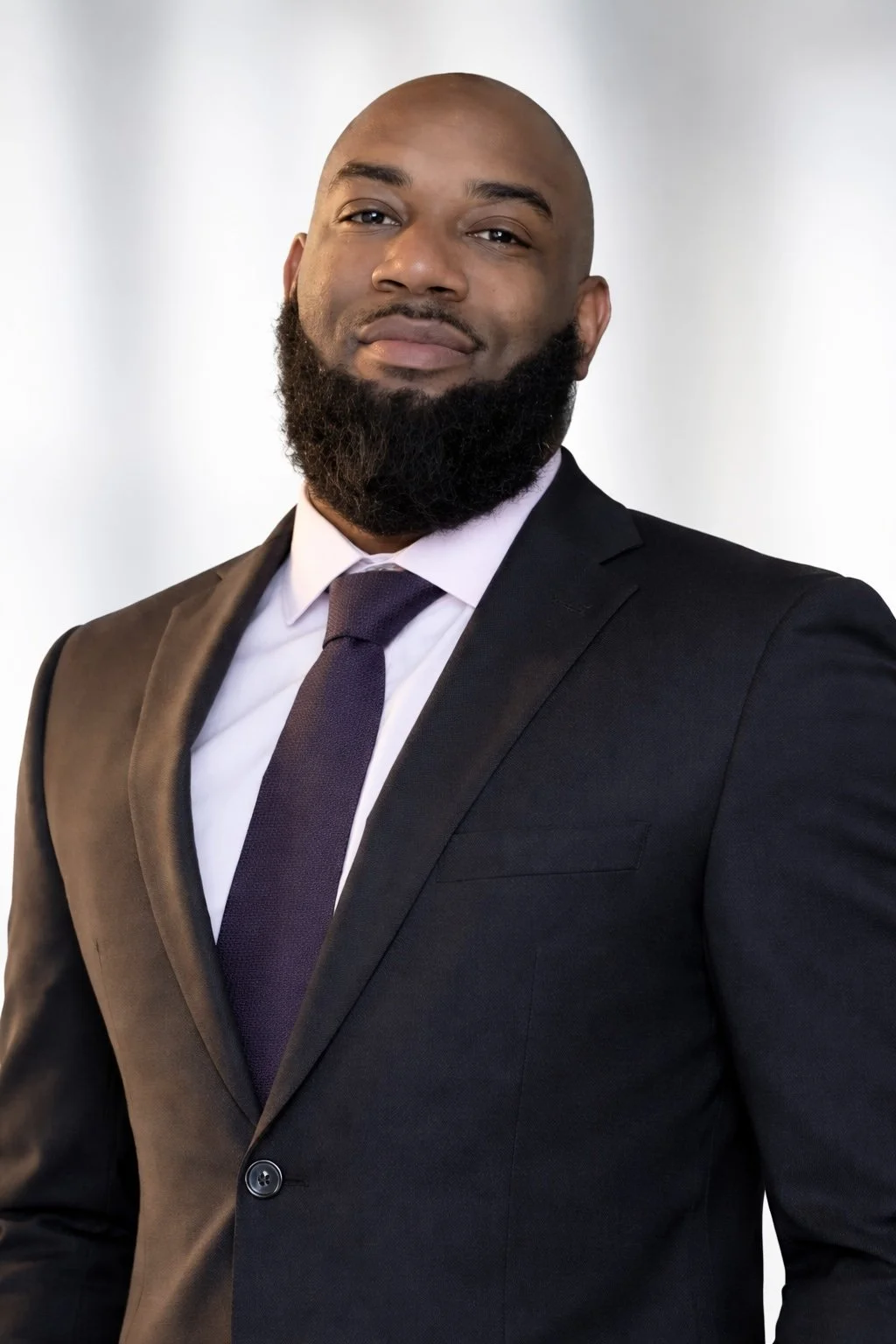 A confident African American man wearing a black suit, light pink shirt, and purple tie, posing against a plain white background.
