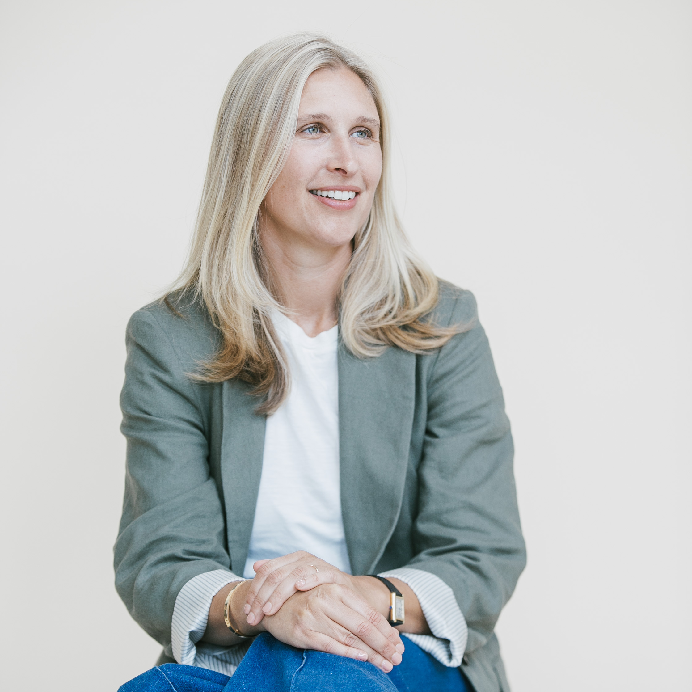 A woman with blonde hair, wearing a green blazer over a white shirt, sitting with her hands folded, smiling and looking to the side.