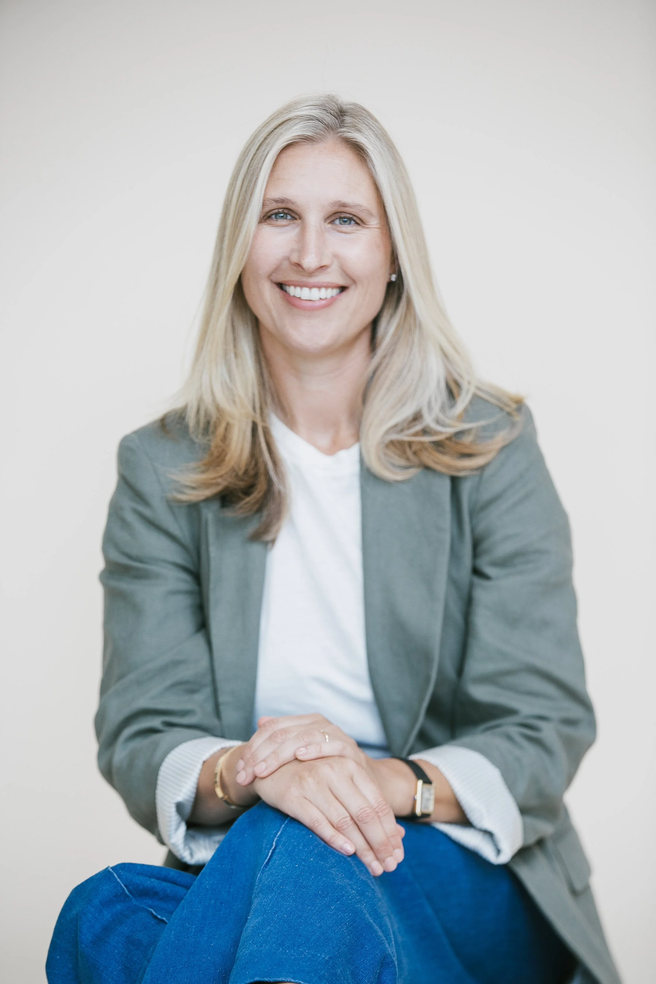 Portrait of a woman with blonde hair, smiling, wearing a gray blazer, white shirt, and blue jeans, sitting with hands clasped.