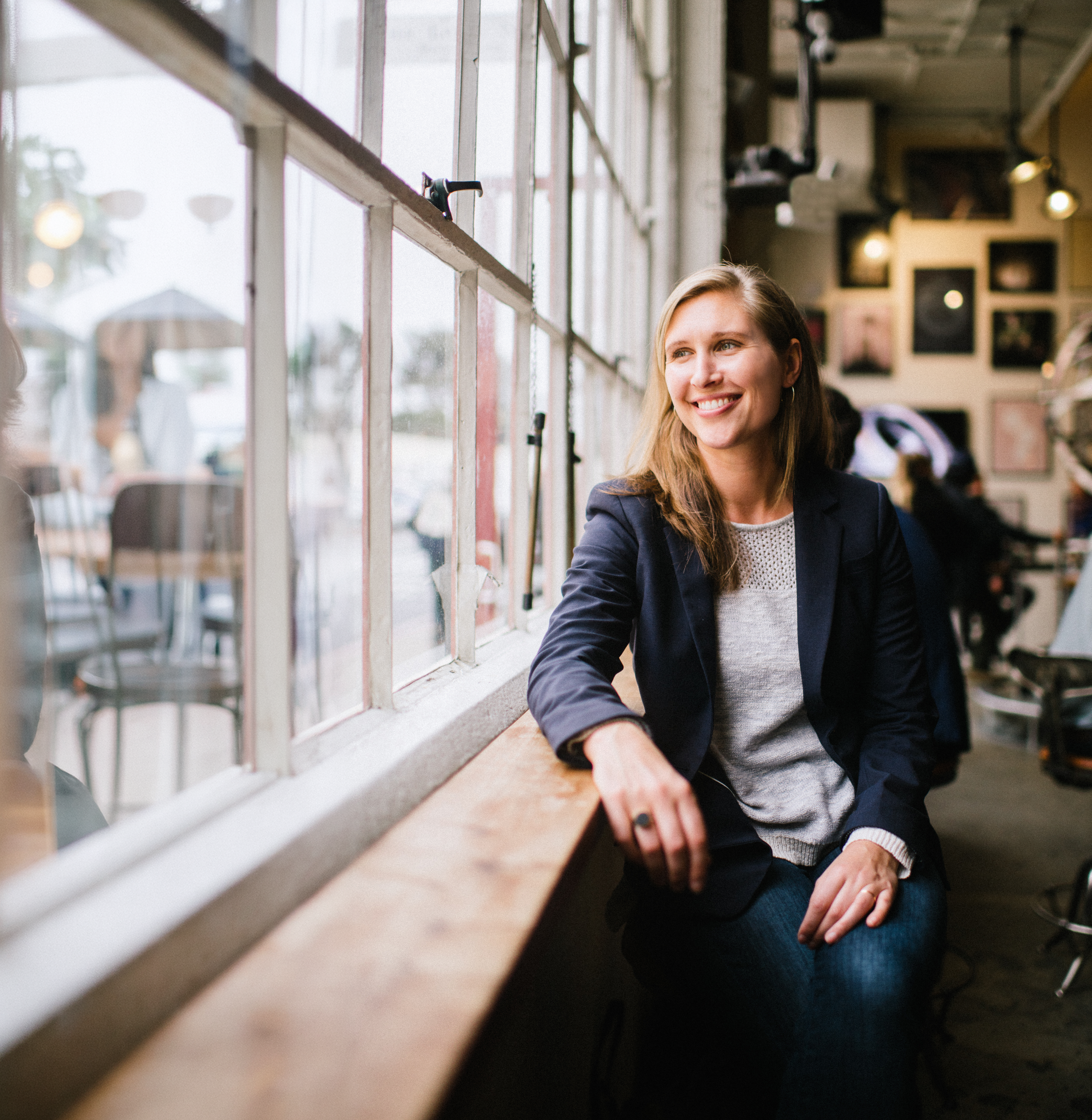 A woman sitting by a large window in a cafe, smiling, wearing a navy blazer over a gray sweater.