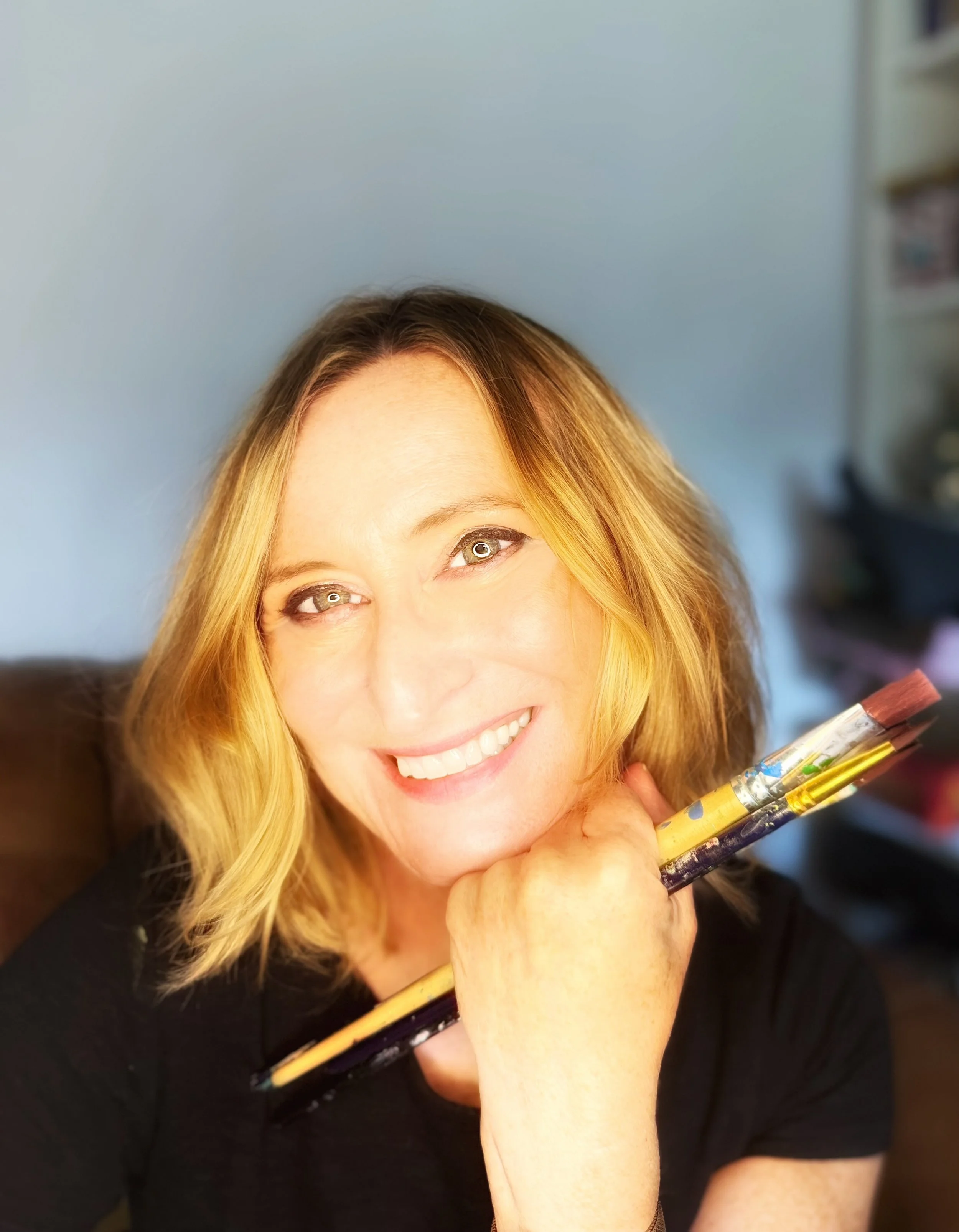 Close-up of Susan Hone Climate artist, smiling, holding a paintbrush, in a room with a blue wall and blurred bookshelf in the background.