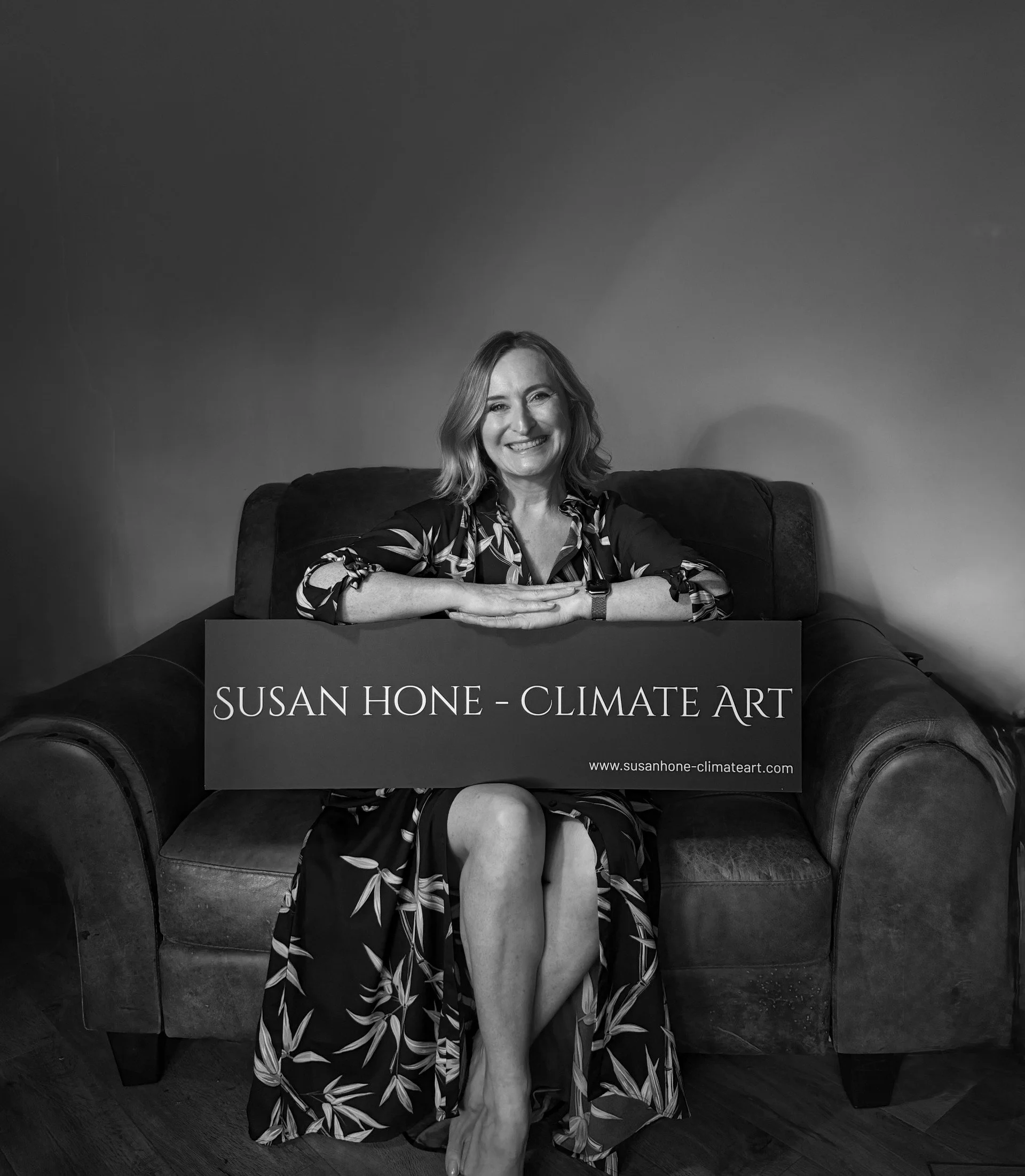 Black and white photo of a woman sitting on a couch, smiling, with a sign reading 'SUSAN HONE - CLIMATE ART' placed in front of her. The woman is dressed in a patterned dress, with her arms resting on the sign, and her legs crossed, showing one leg partly uncovered.