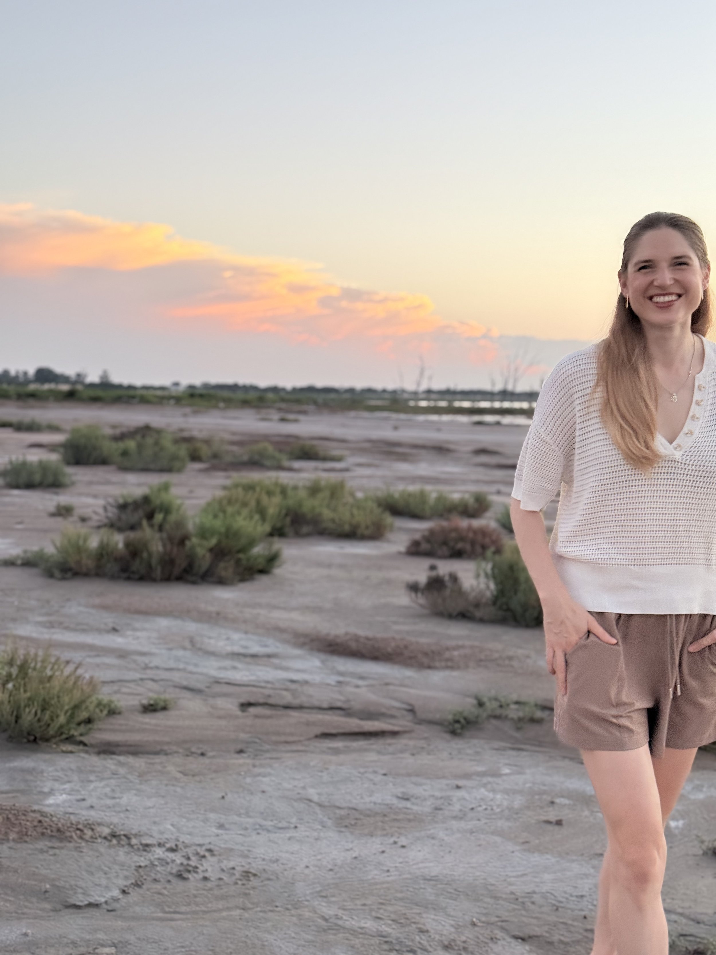 A woman with long hair, wearing a white top and brown shorts, smiling while standing outdoors in a desert or dry landscape during sunset or sunrise.