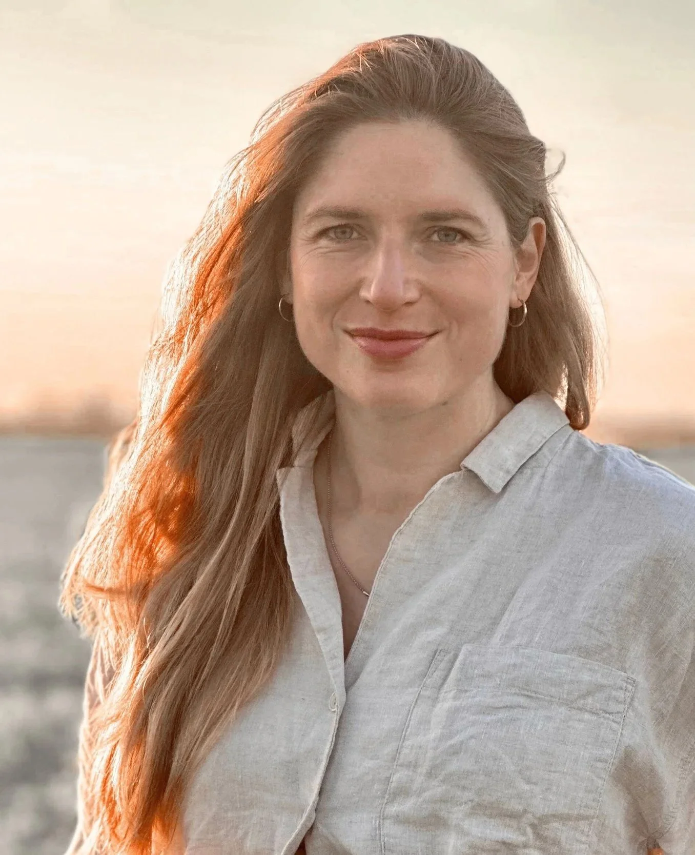 A woman with long, wavy, reddish-brown hair standing outdoors during sunset, wearing a light-colored button-up shirt, smiling softly at the camera.