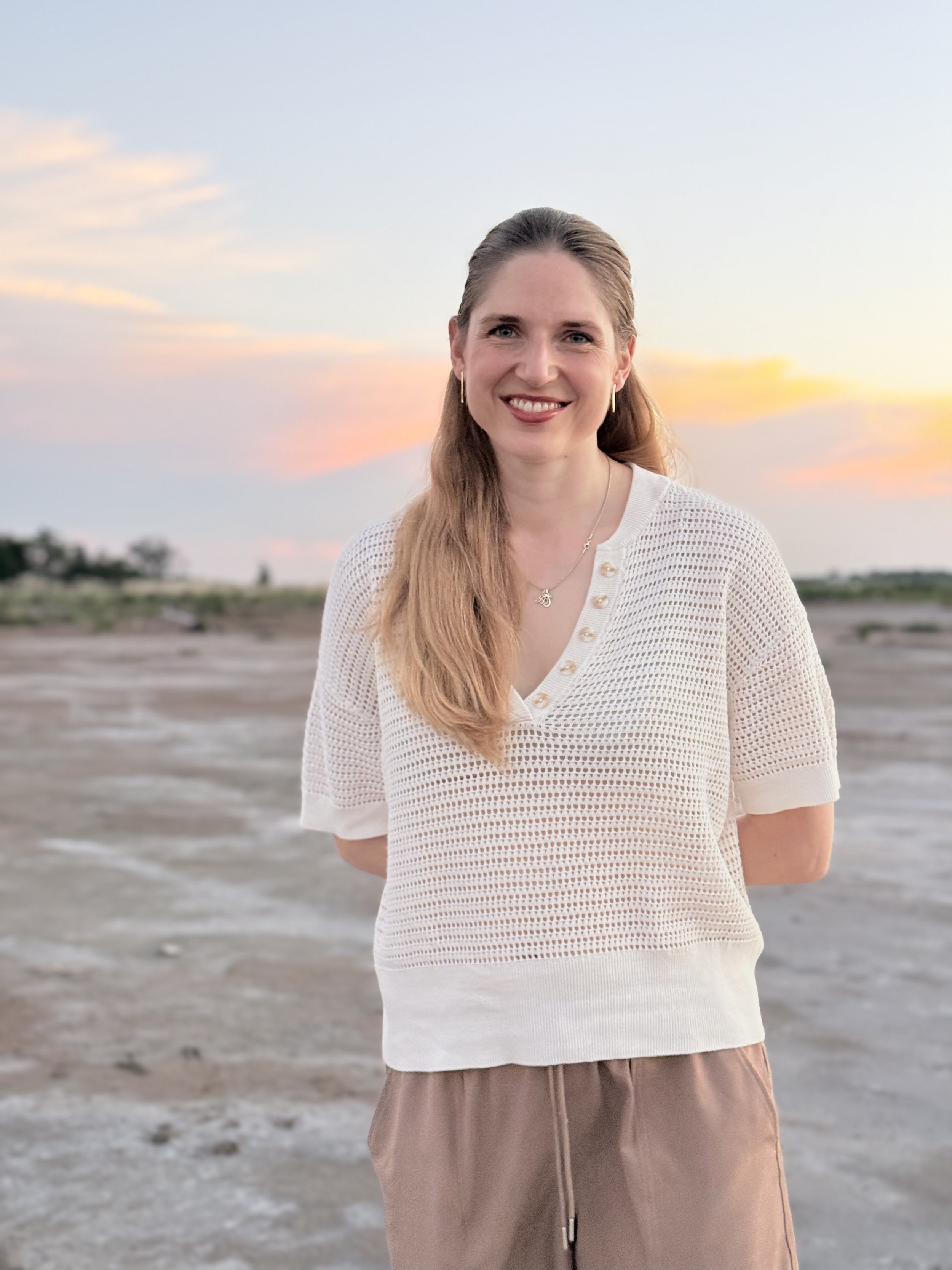 A woman with long, light brown hair smiling outdoors during sunset, wearing a white knitted short sleeve top, earrings, and a necklace, with a blurred background of sky and open land.