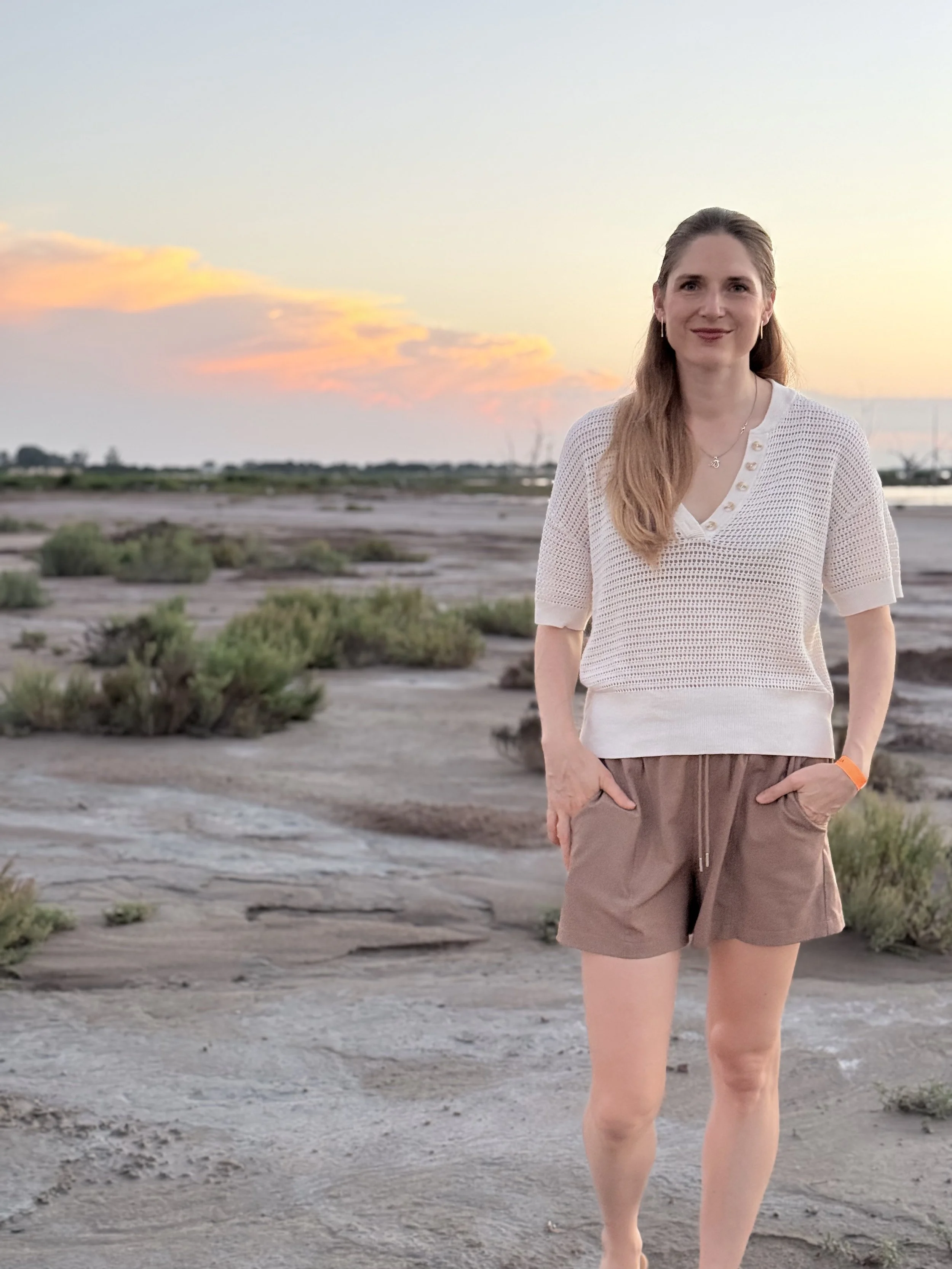 A woman with long hair smiling, standing outdoors on a rocky and sandy terrain with sparse vegetation, during sunset or sunrise, with clouds in the sky.