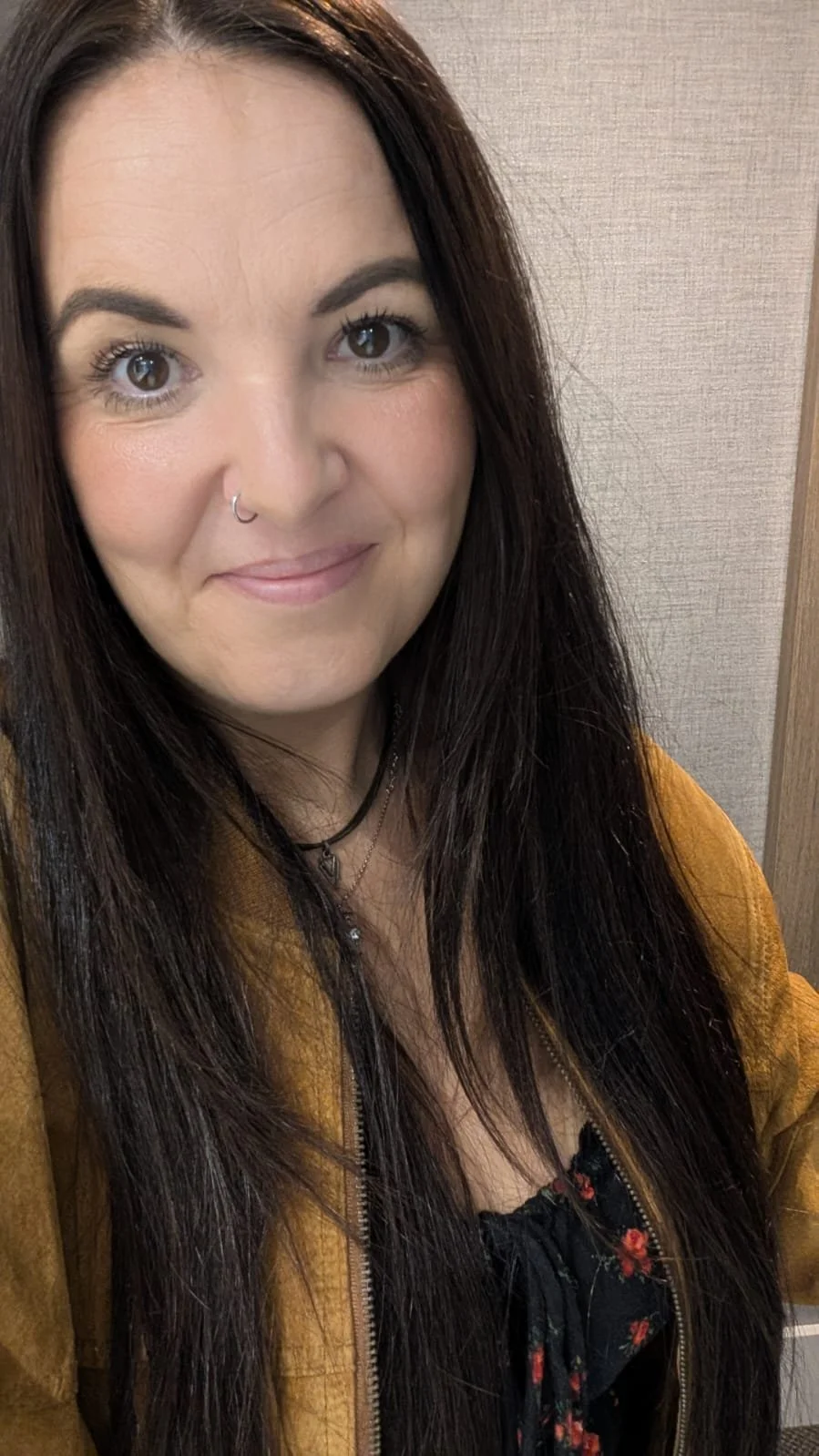 A woman with long dark hair, light skin, and brown eyes, smiling, wearing a nose ring, a necklace, a black floral top, and a mustard-colored jacket, standing against a beige wall.