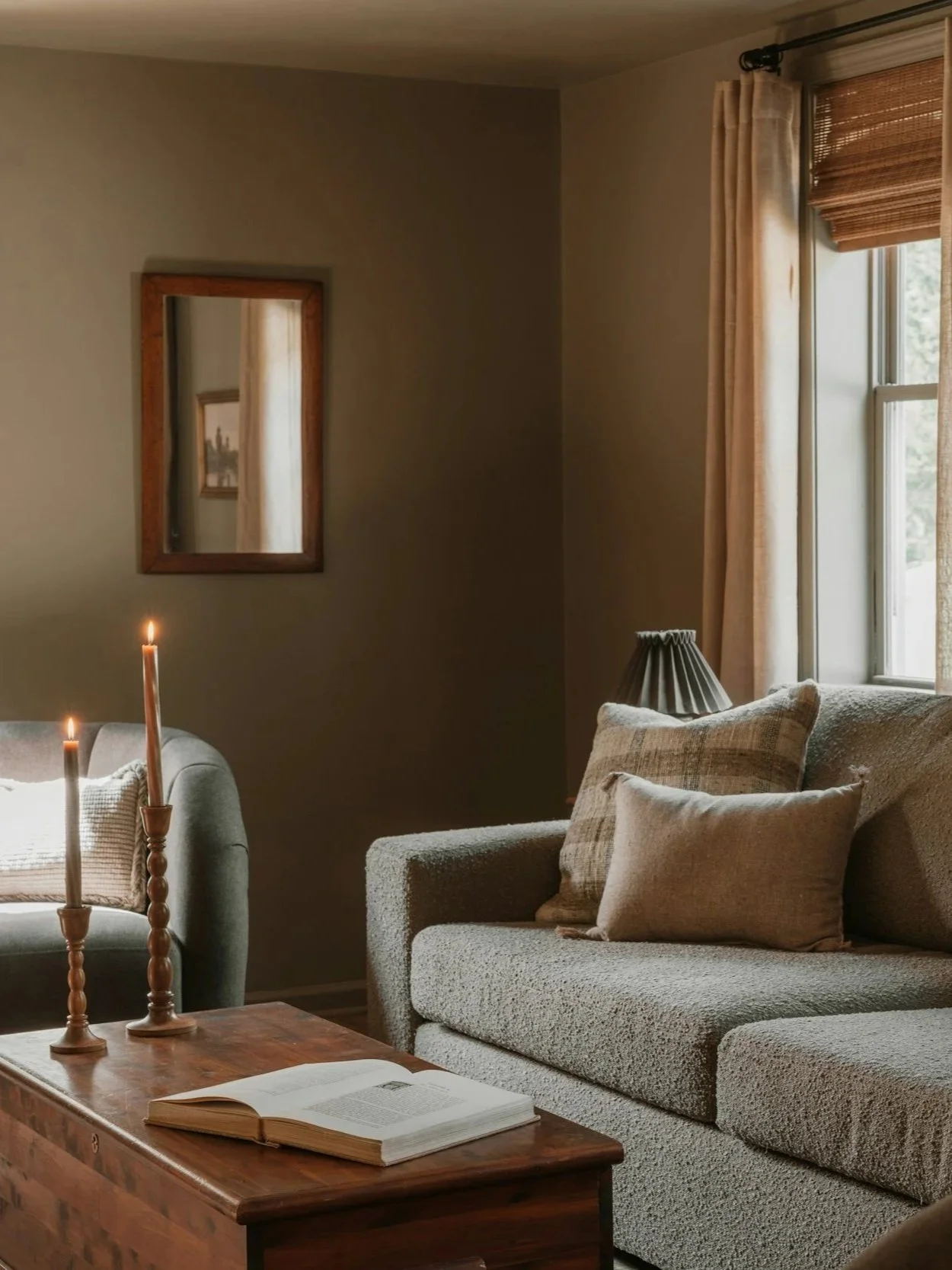 Cozy living room with beige sofas, pillows, a wooden coffee table with an open book, candles, and a window with curtains.