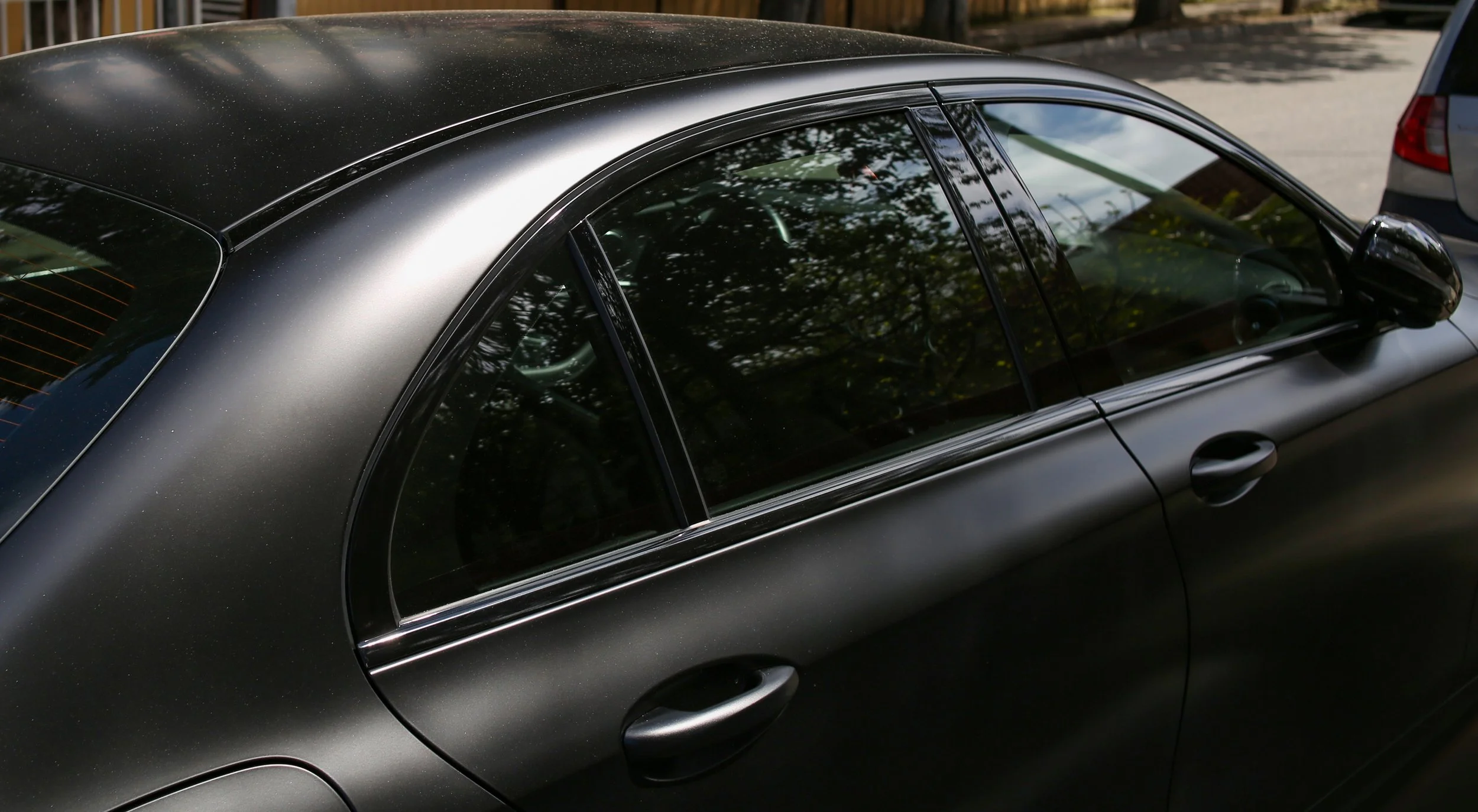 Close-up of a black sedan car parked outdoors, showing the side windows, door handles, and part of the roof with reflections of trees.