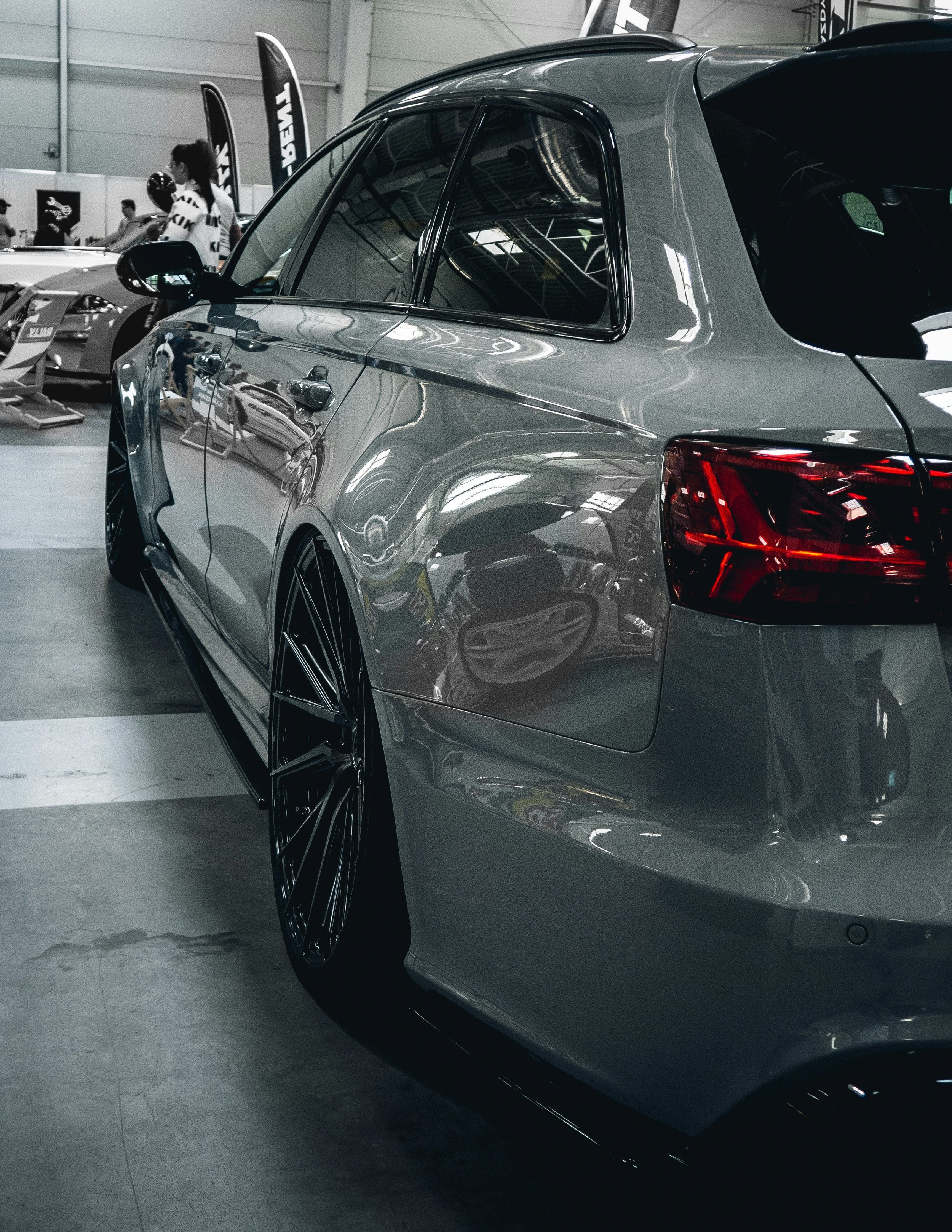 A sleek gray car with black rims displayed at an indoor auto show, with reflective surfaces and people in the background.