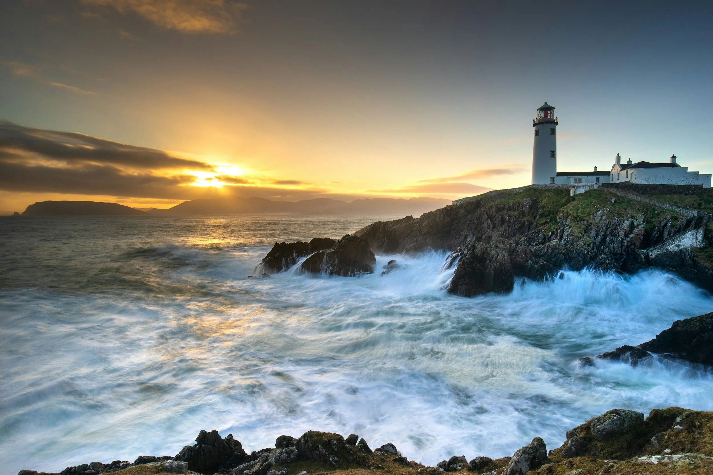 Sunset over the ocean with waves crashing against rocky coast and a white lighthouse on a grassy hill.
