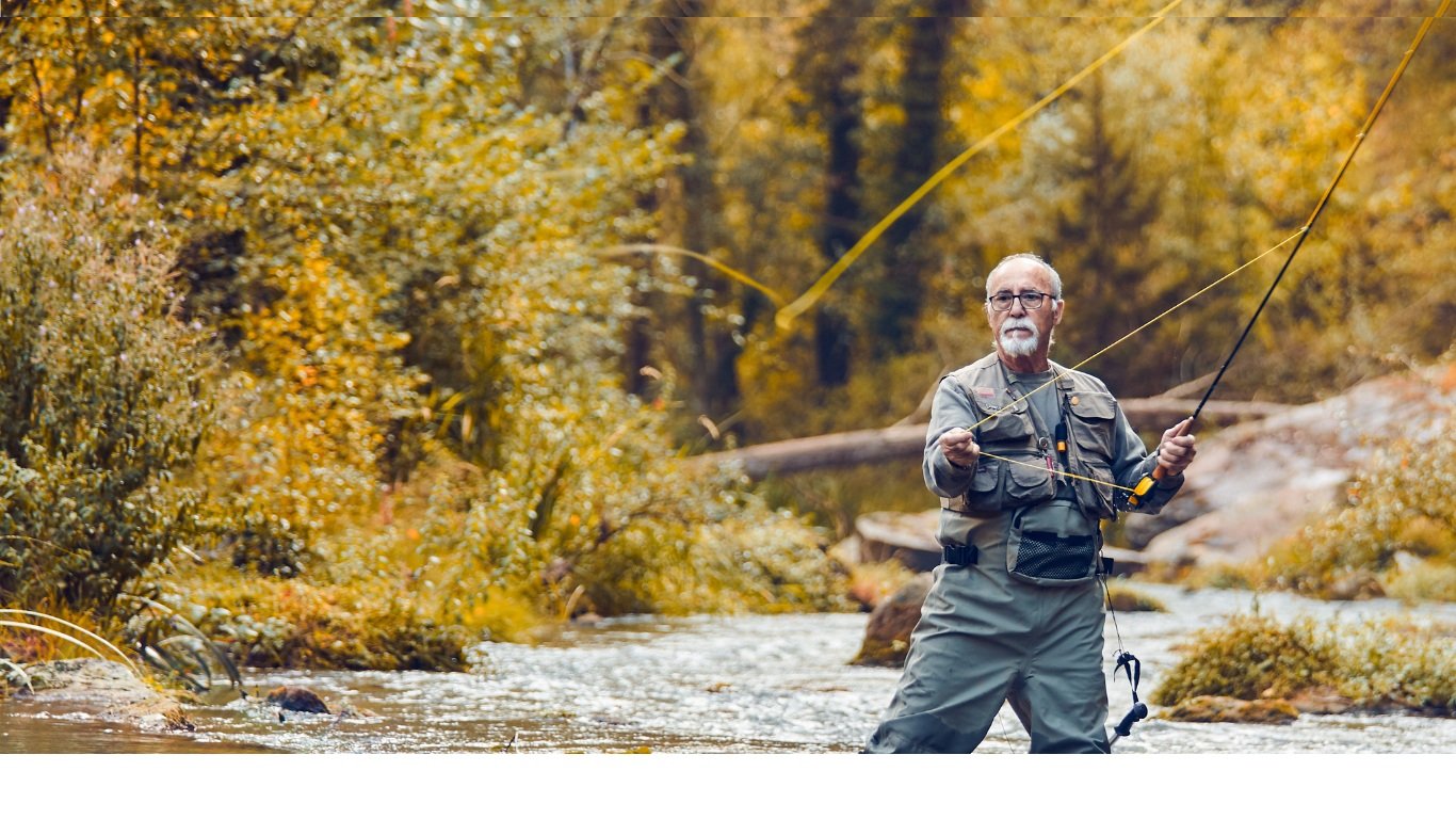 An older man fishing in a river surrounded by autumn-colored trees.